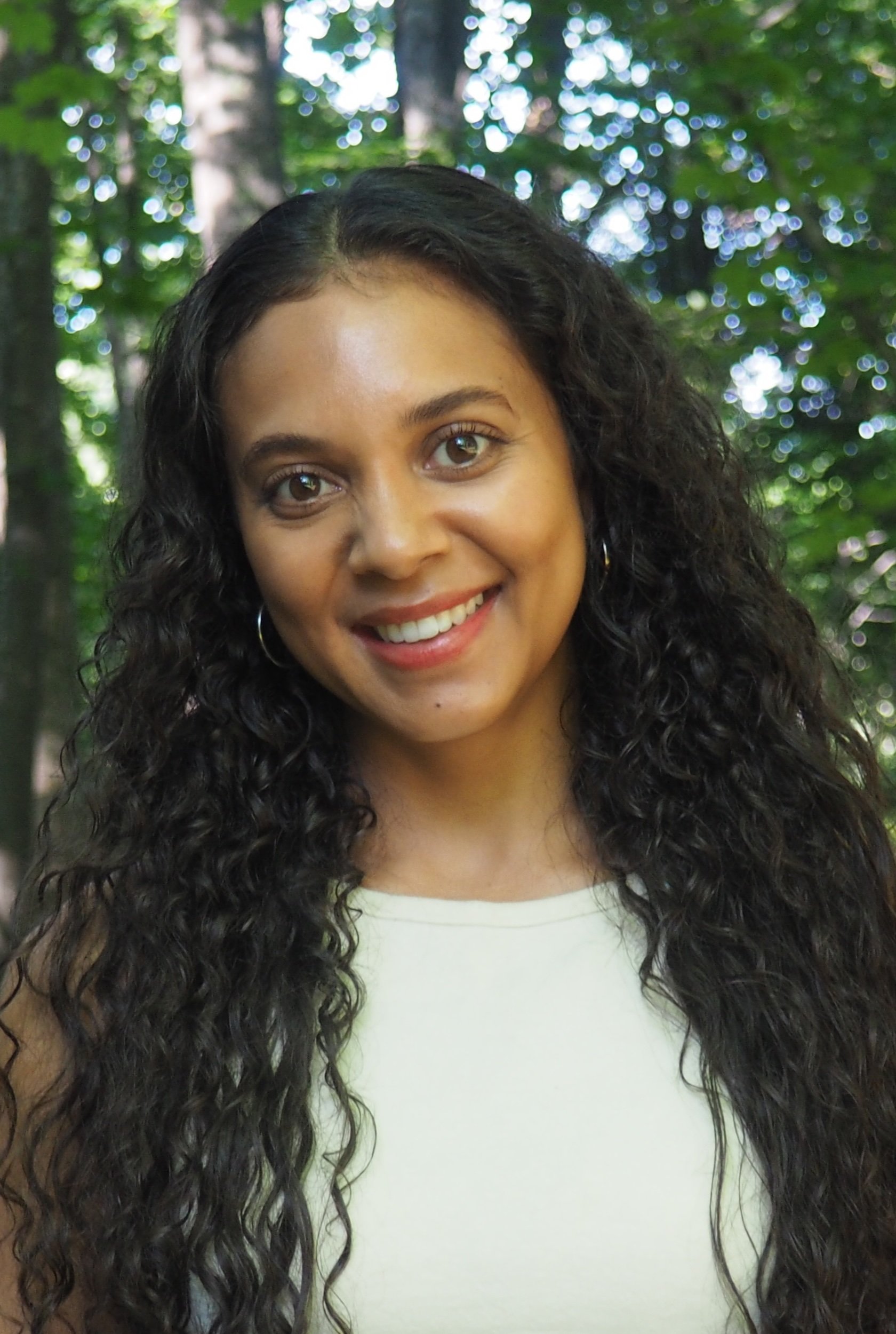 merinda valley: headshot of a smiling woman with long curly black hair, wearing a white top and hoop earrings, standing outdoors with a background of green trees and dappled sunlight.