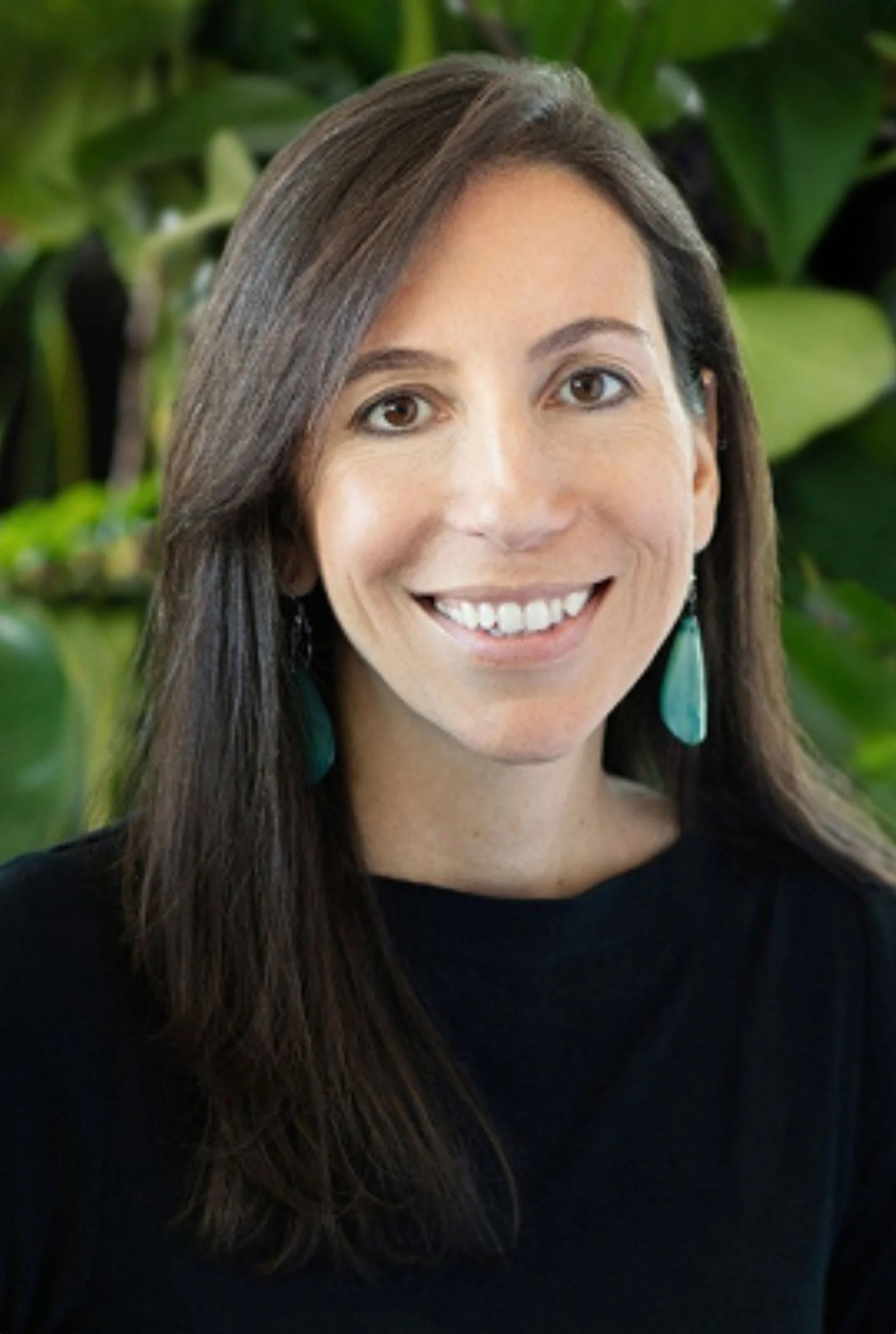 sandrina da cruz: headshot of a smiling woman with long brown hair, wearing turquoise earrings and a black top, with green leaves background.