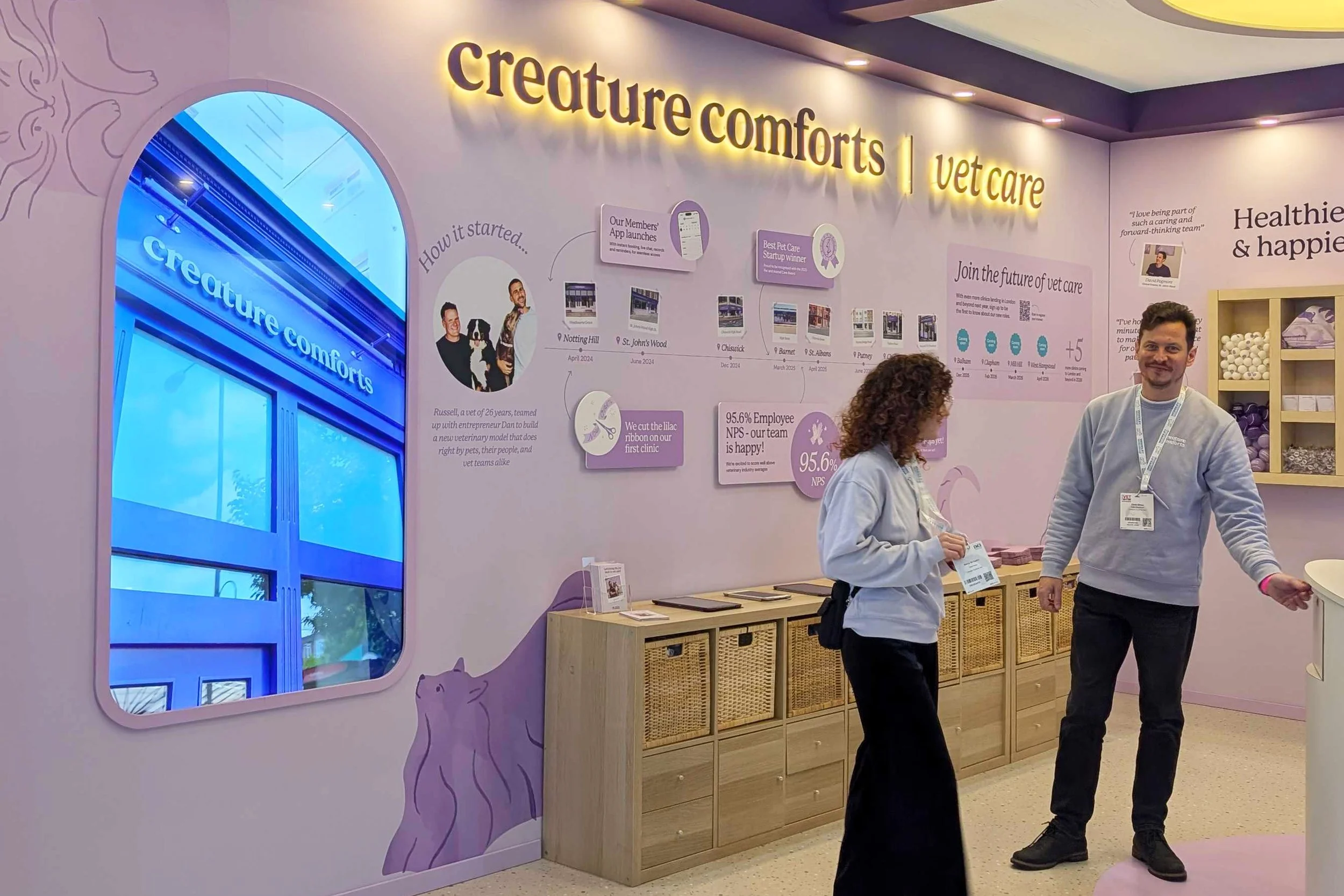 Interior of a veterinary clinic called Creature Comforts Vet Care with two staff members talking. The wall has informational graphics about the clinic's history and achievements, with a large window displaying the clinic's name.