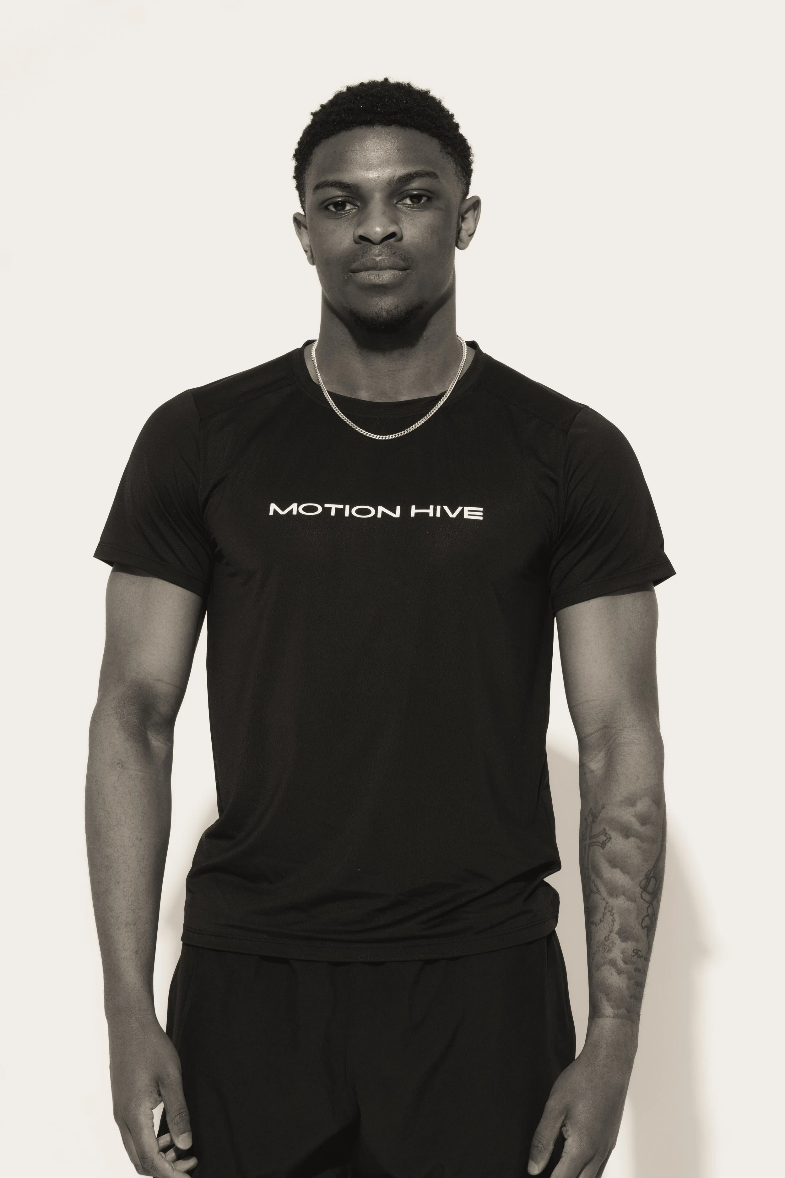 A young man with short curly hair, wearing a black t-shirt with the words 'MOTION HIVE' and a small silver necklace, stands against a plain white background.