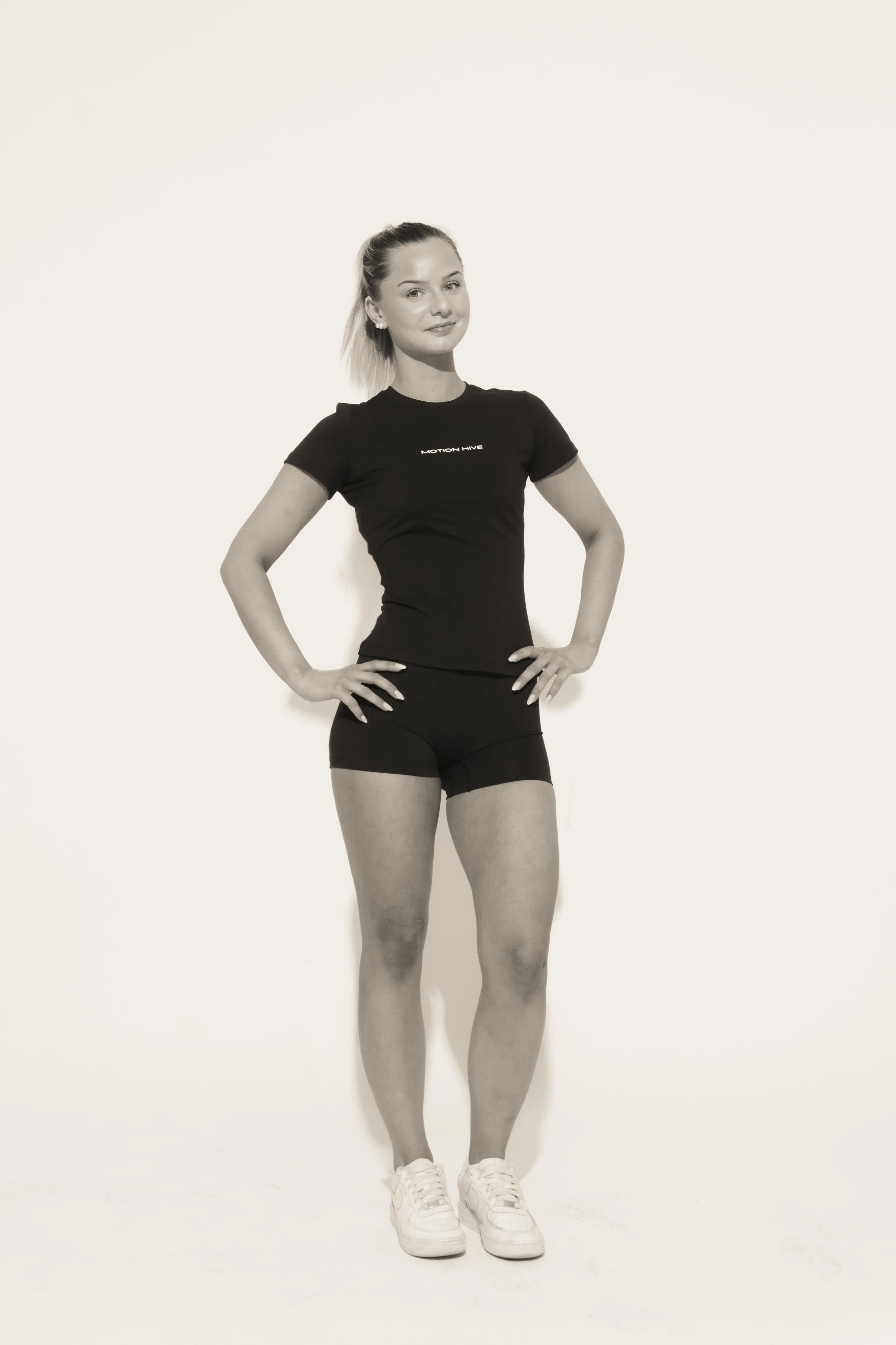 A young woman in sportswear stands with hands on hips against a plain white background, smiling slightly.