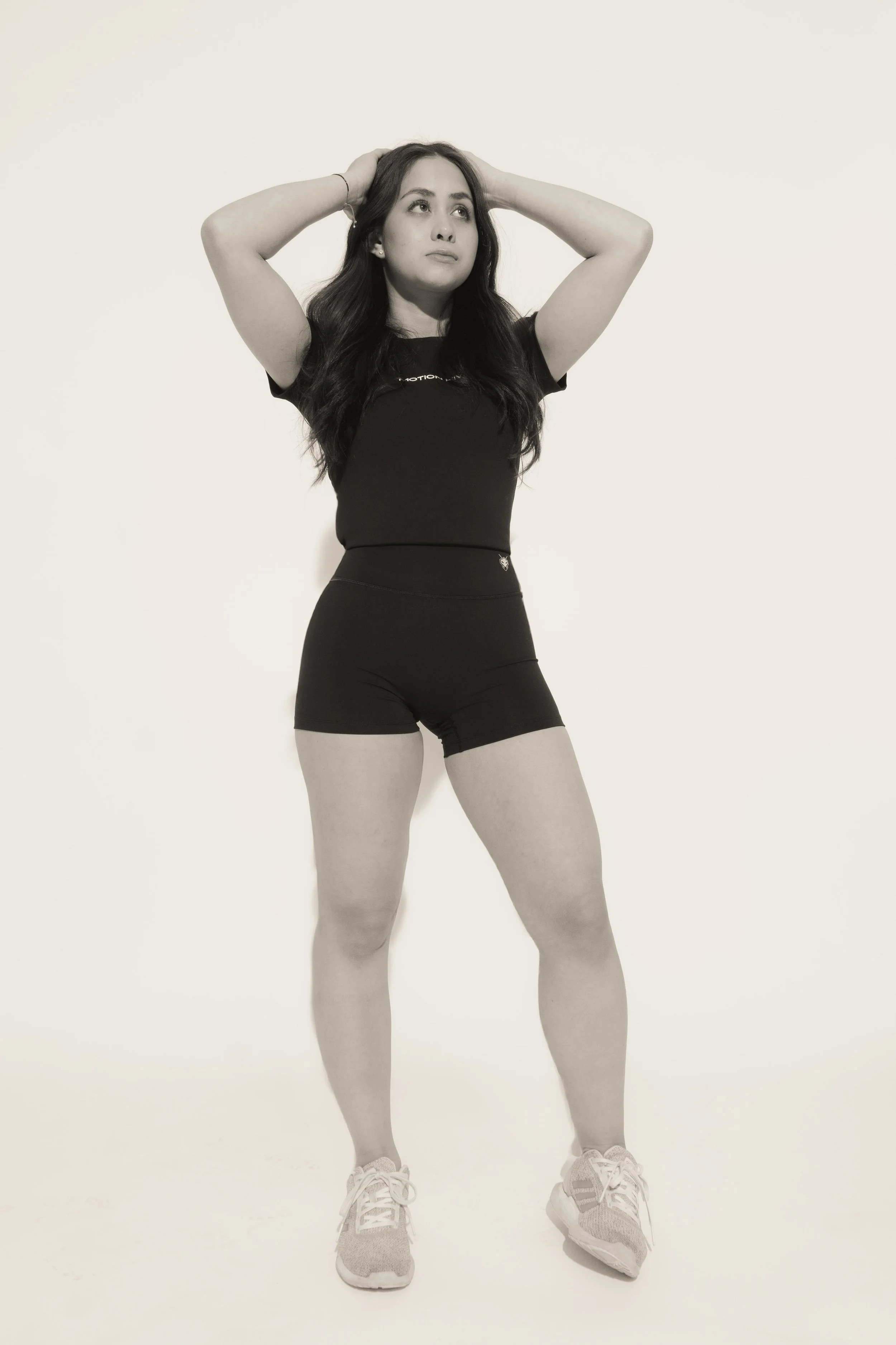 Young woman standing against a plain background, with her hands behind her head, wearing a black athletic T-shirt, black shorts, and running shoes.