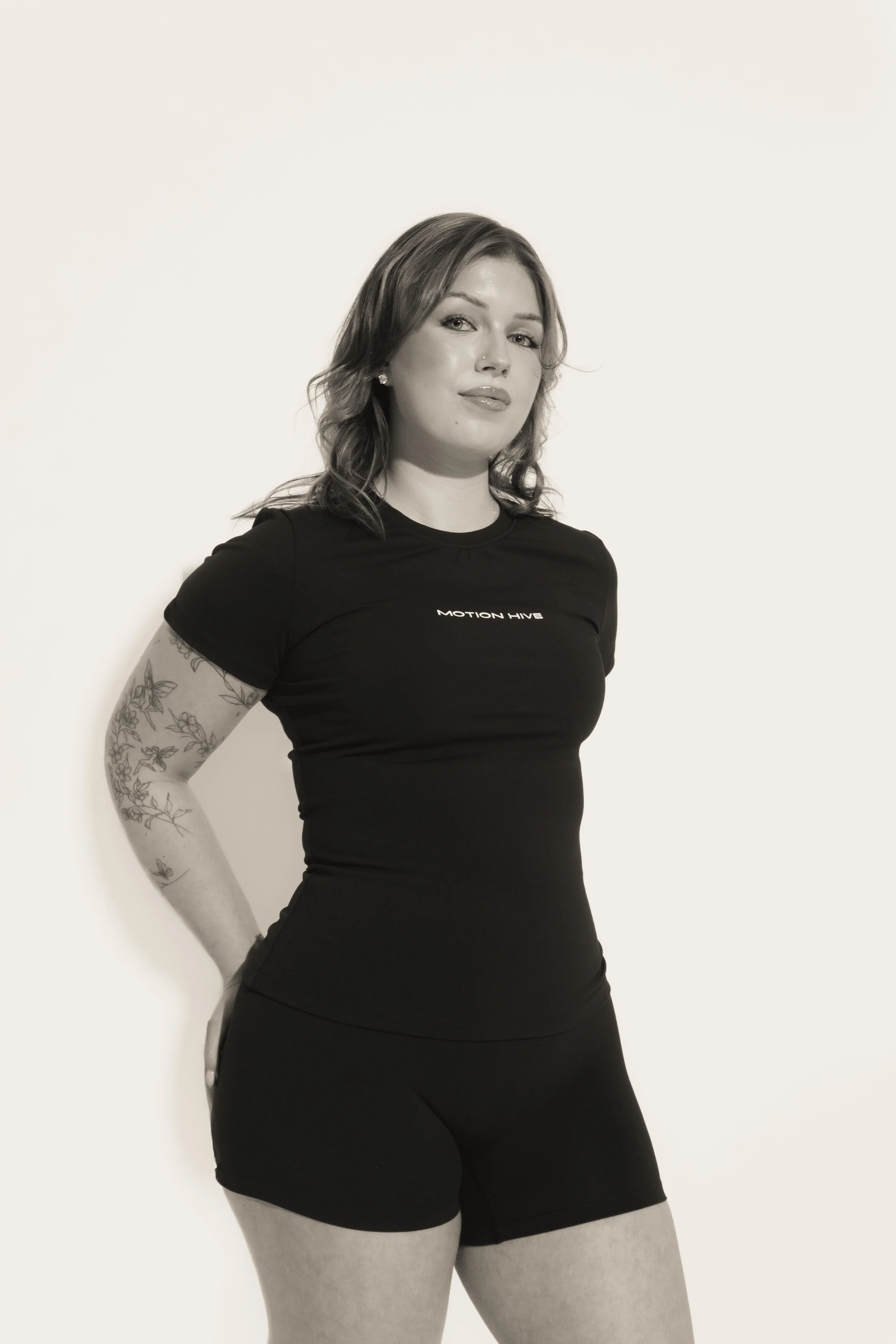 Black and white photo of a woman with shoulder-length wavy hair, wearing a fitted black T-shirt with 'Motion Hive' written on it, standing against a plain light background, with her arms behind her back and looking confidently at the camera.