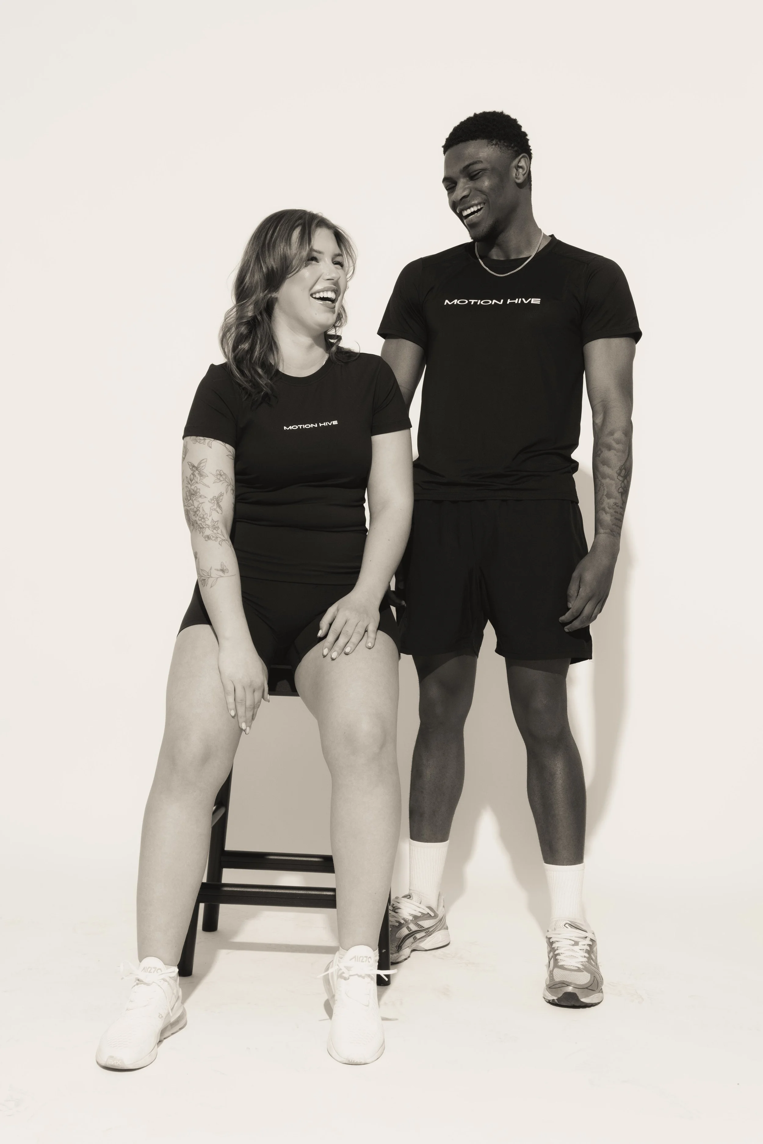 Black and white photo of two people in athletic clothing, one sitting on a chair with legs apart and the other standing, both smiling and looking at each other, against a plain background.