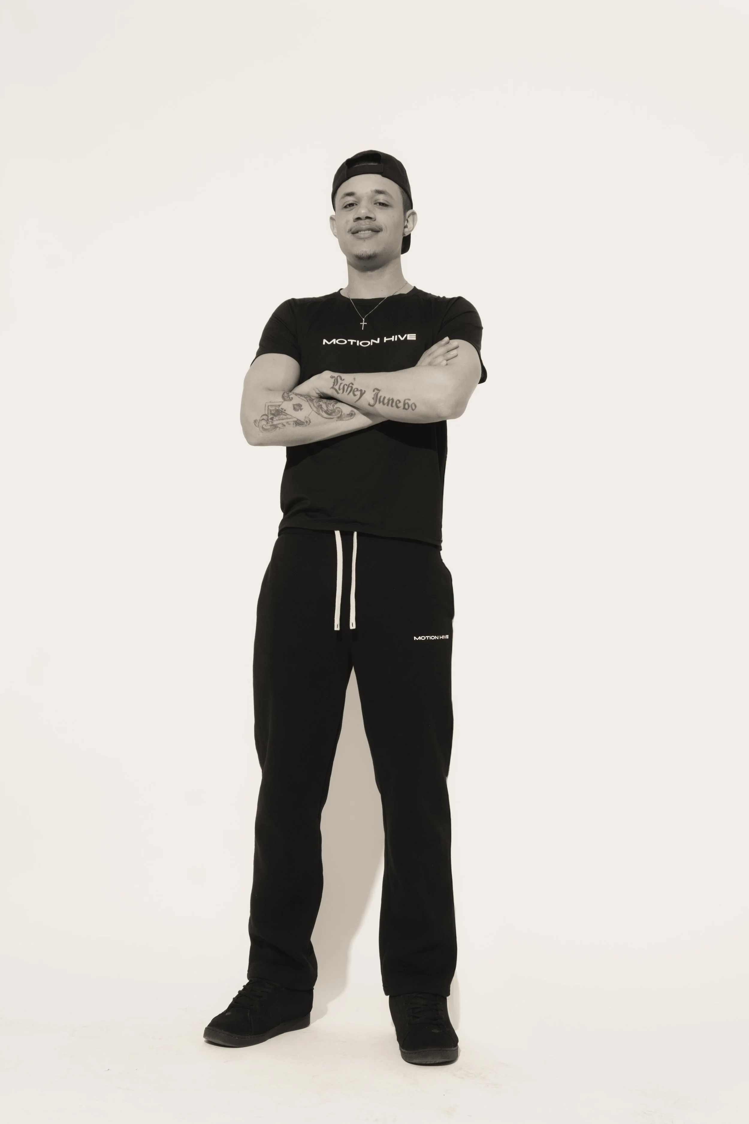 A young man standing with arms crossed, wearing a black t-shirt, black sweatpants, black shoes, a backwards black cap, a necklace with a cross pendant, and has tattoos on his arms, against a plain white background.