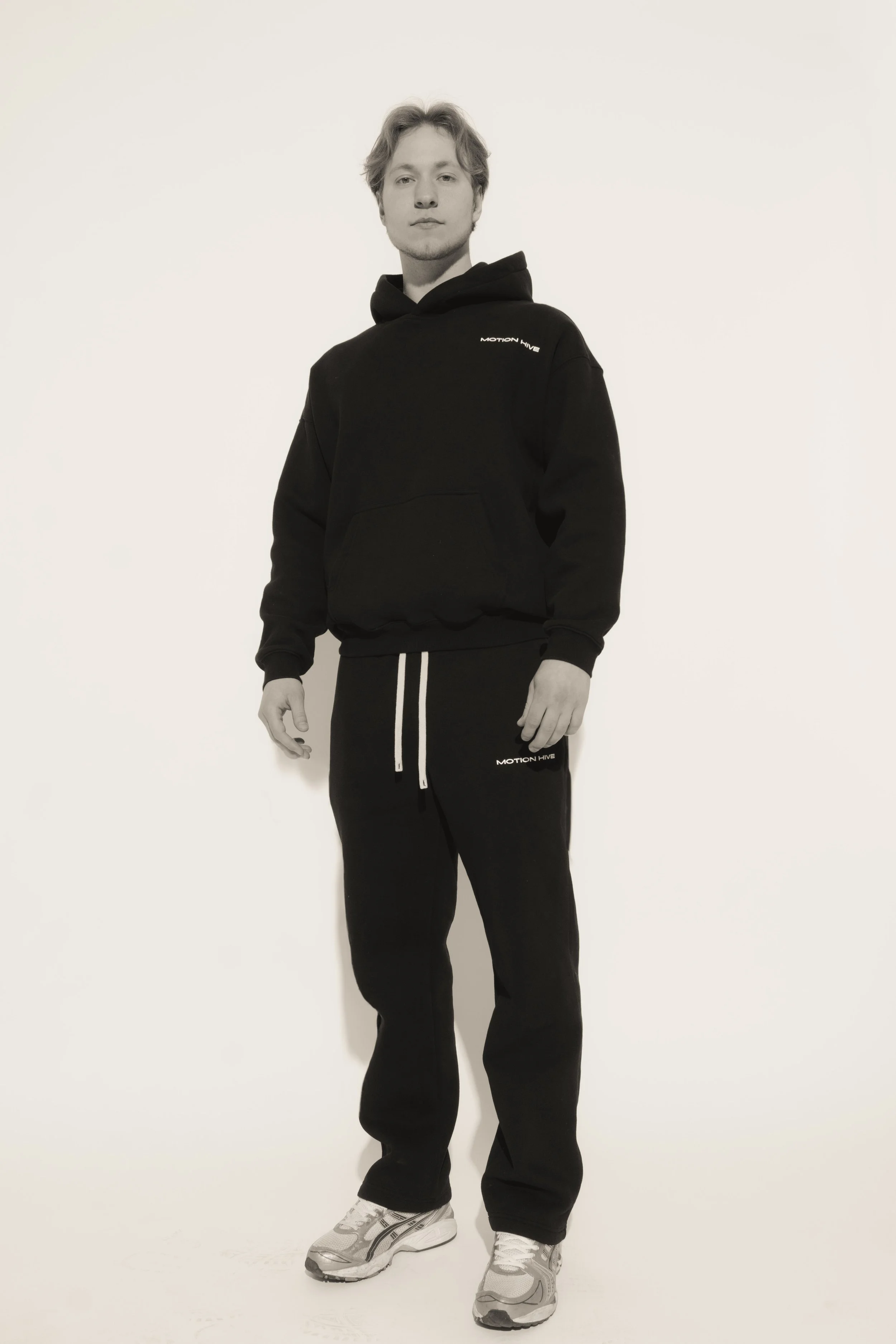 A young man standing against a plain background, wearing a black hoodie with 'MOTION HIVE' text on it, black sweatpants, and gray sneakers.