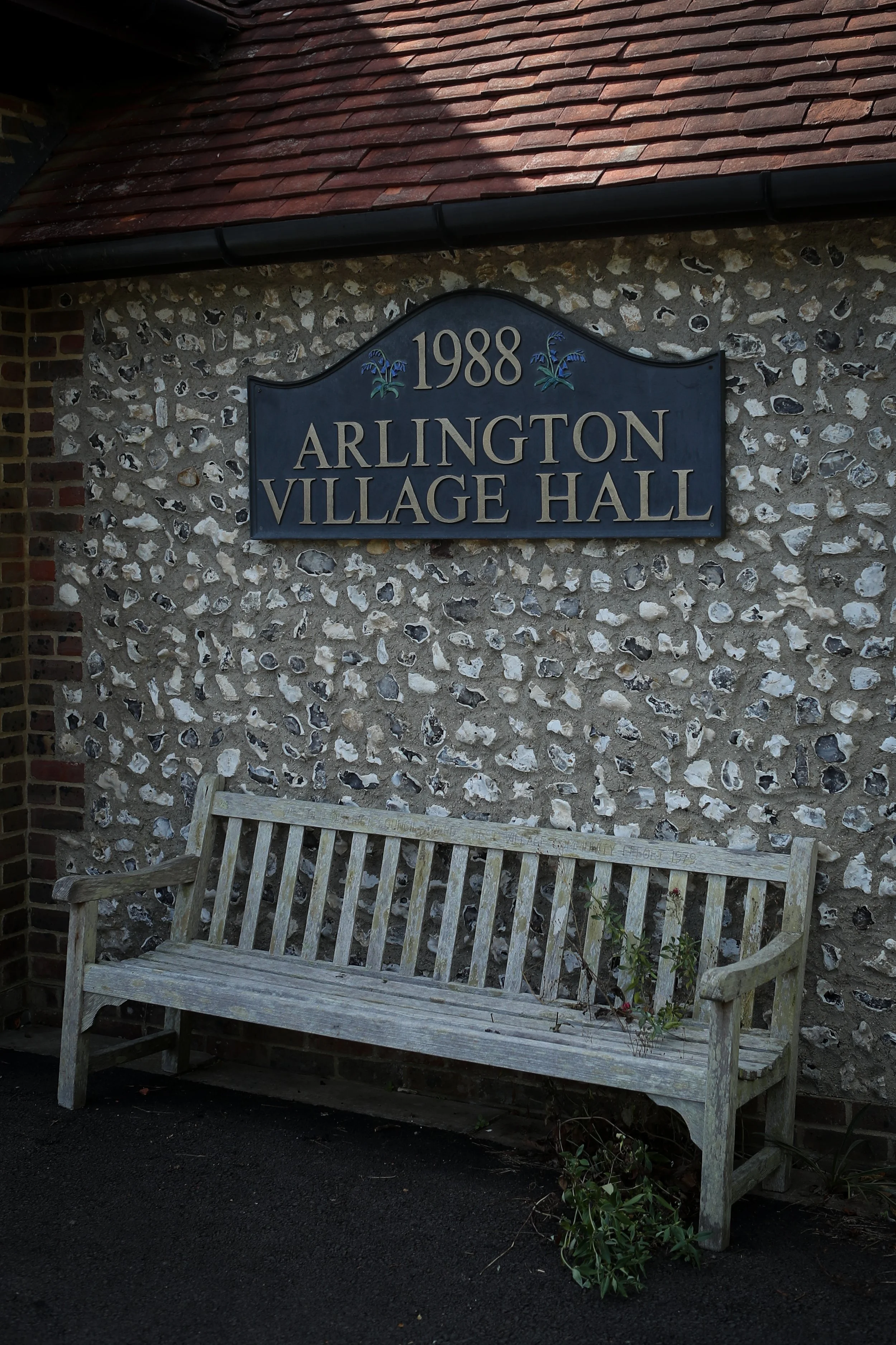 A weathered wooden bench in front of a stone wall with a sign that reads '1988 Arlington Village Hall.'