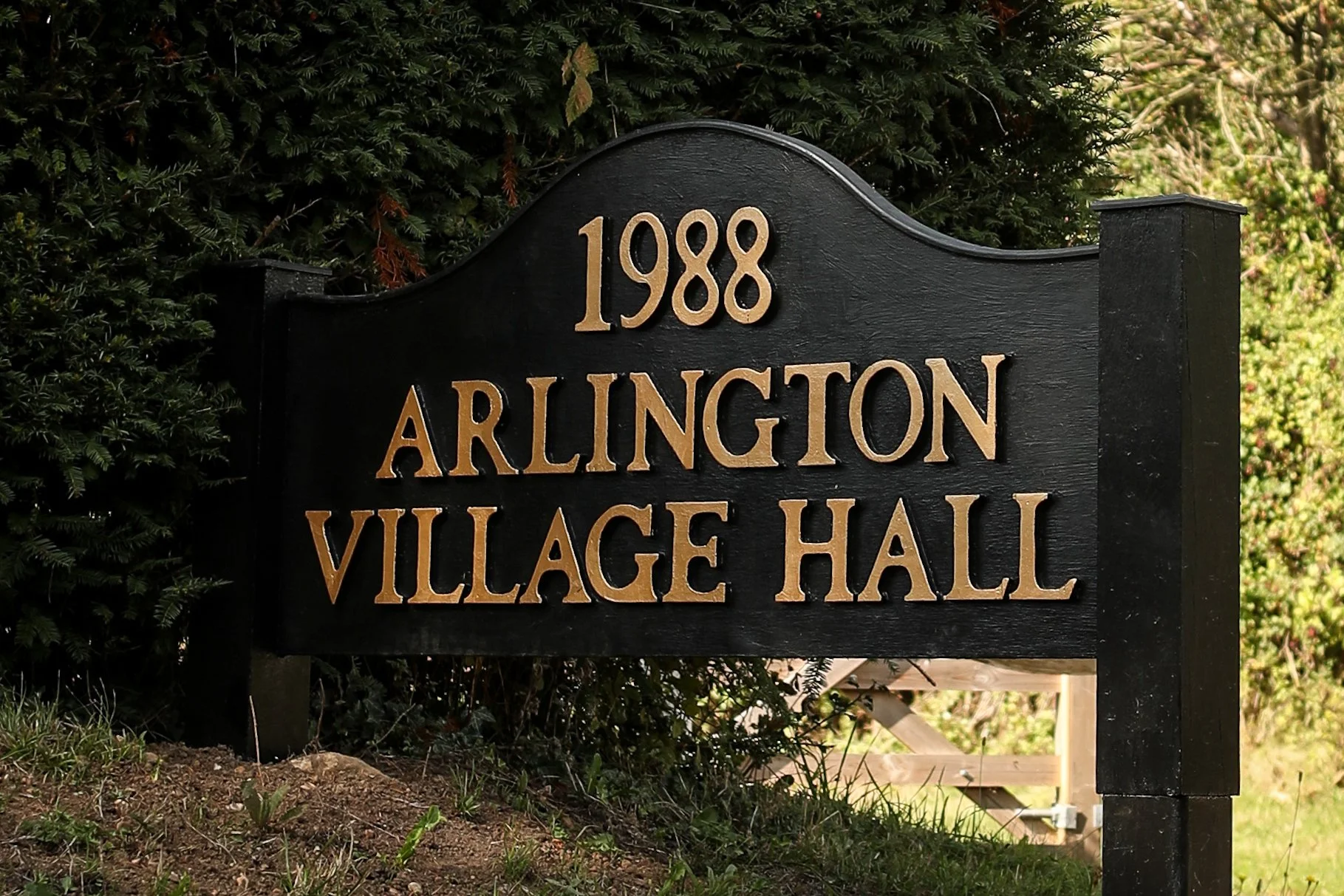 A black wooden sign with gold lettering reads '1988 Arlington Village Hall', situated outdoors among greenery.