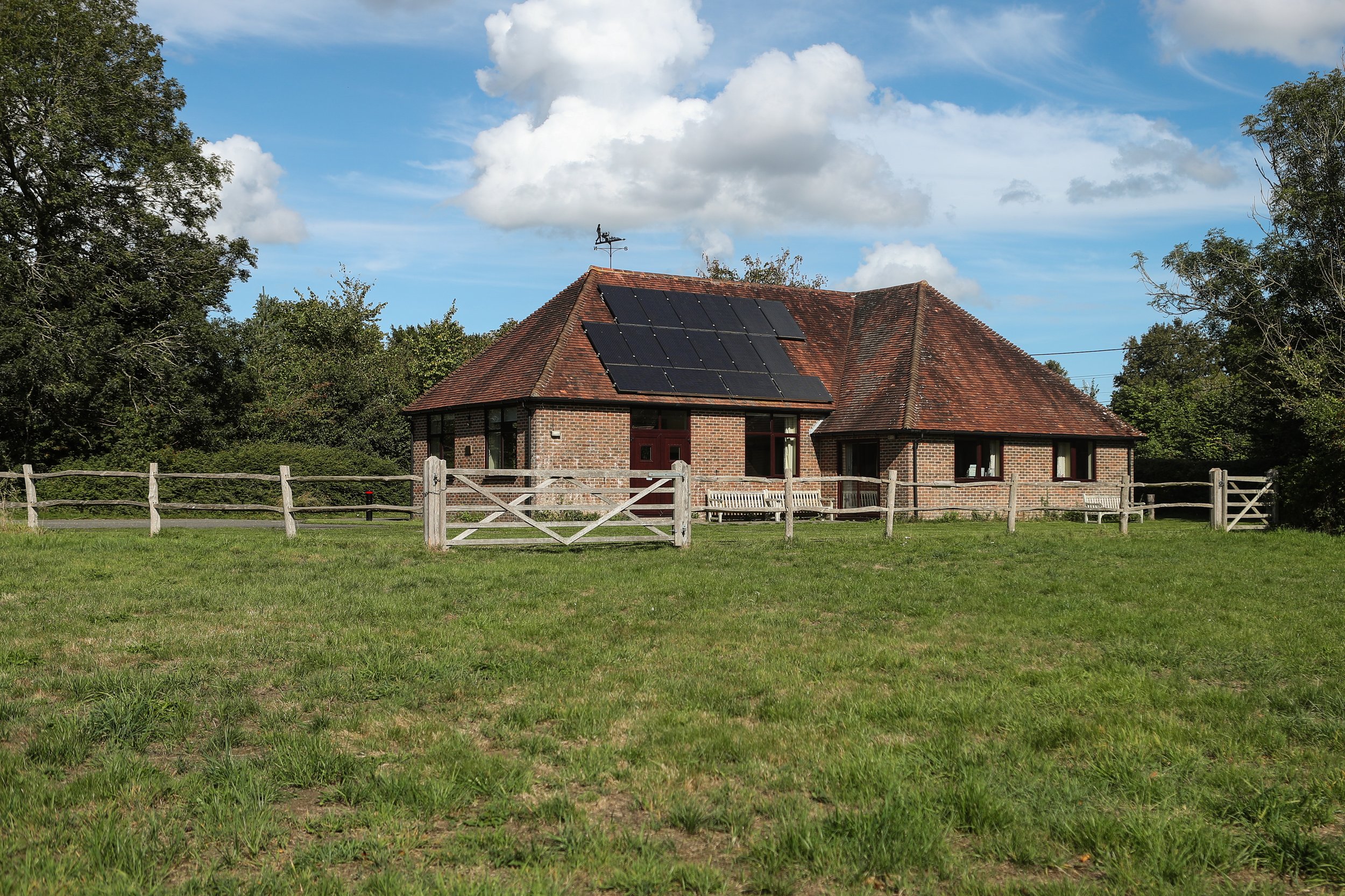 A brick house with solar panels on the roof, surrounded by green grass and trees under a partly cloudy sky.