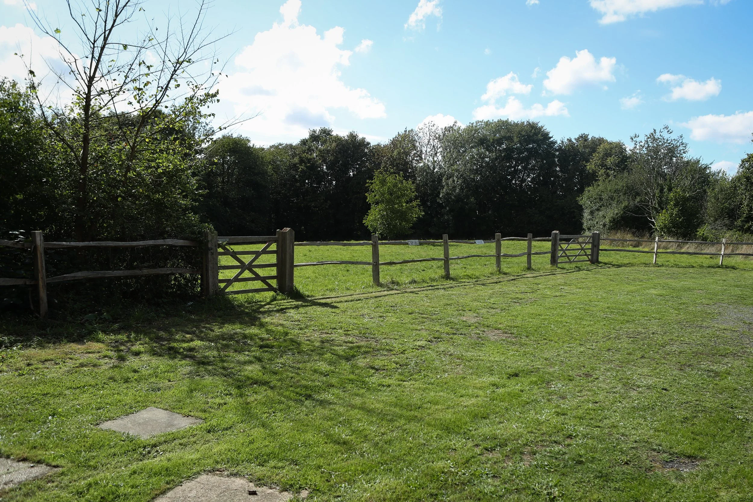 A grassy field enclosed by a wooden fence with small signs, surrounded by trees under a partly cloudy sky.