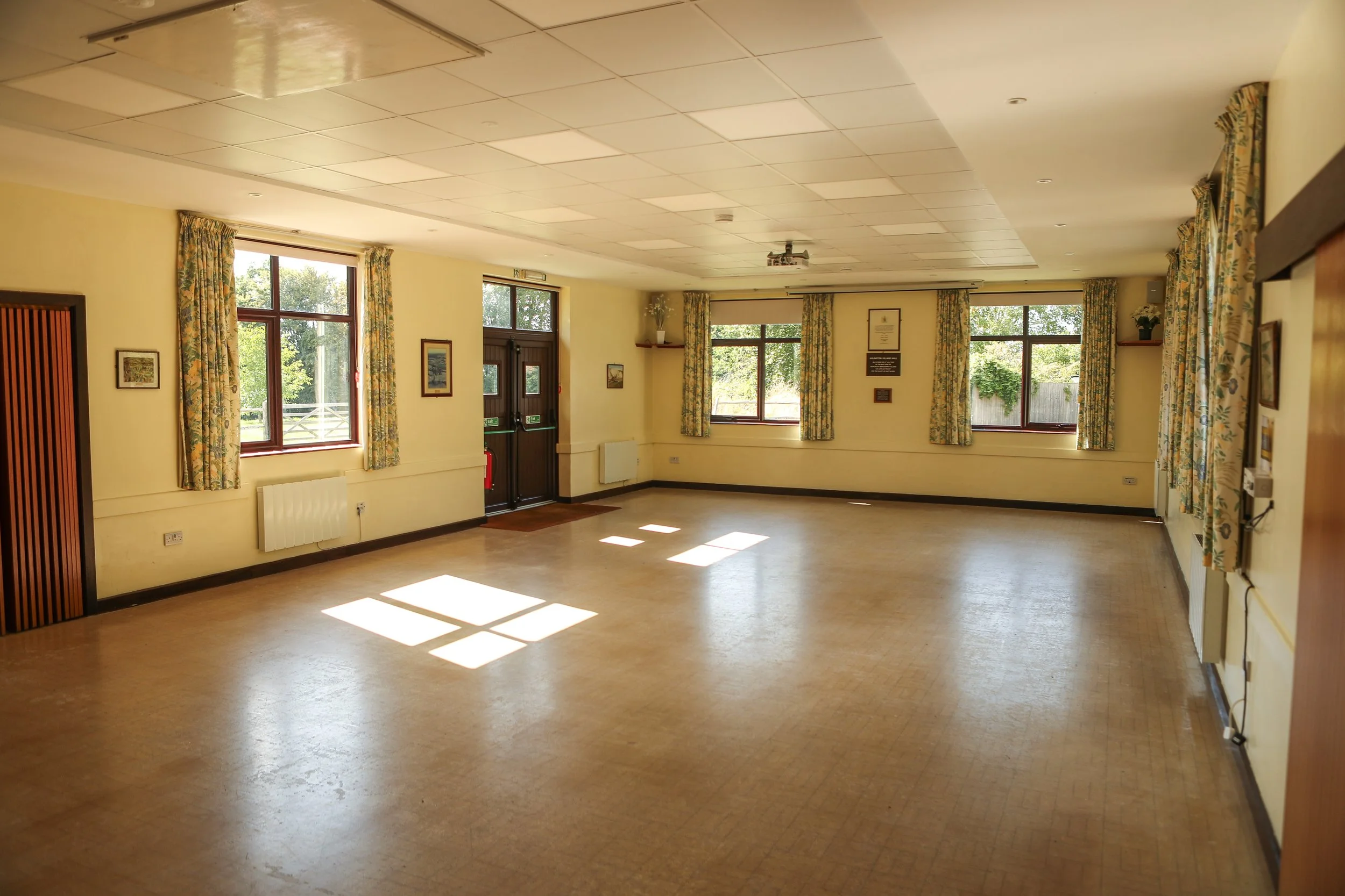 Empty event hall with beige walls, large windows with floral curtains, and a wooden entrance door. Sunlight streams through the windows, casting shadows on the beige tiled floor. Framed pictures hang on the walls.