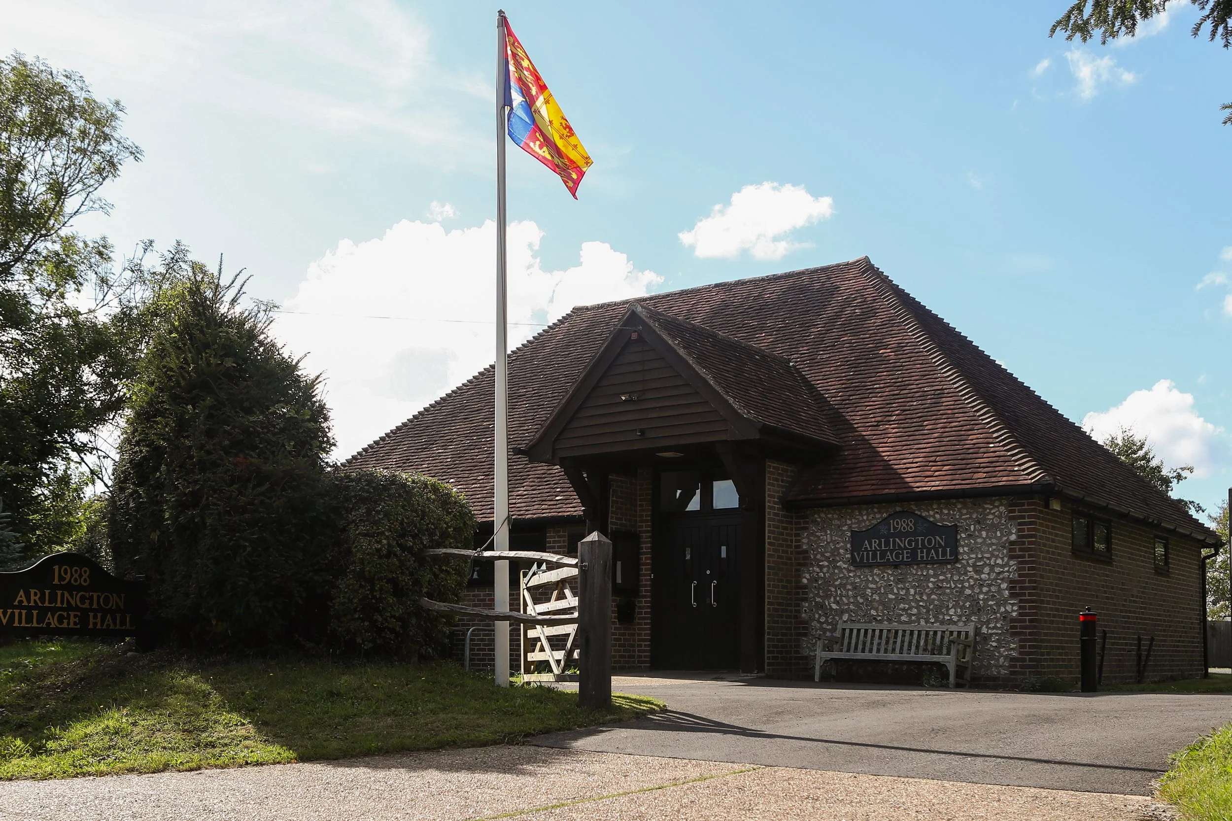 Arlington Village Hall, a brick building with a shingled roof, a small entrance with a gabled porch, a flagpole with a colorful flag, a bench, and a sign indicating it was built in 1988.