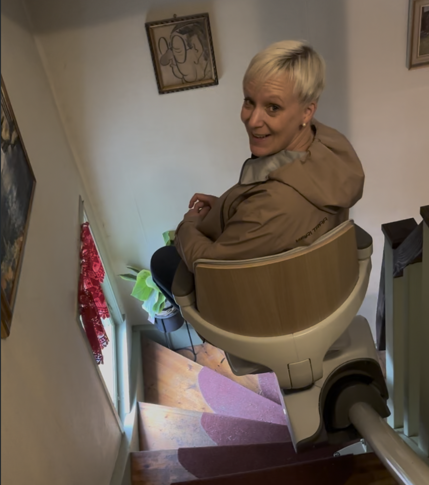 Ergoterapeut fra Ingen Hindring sitter i en stolheis som kjører langs skinne i en smal innendørs svingtrapp med tretrinn.