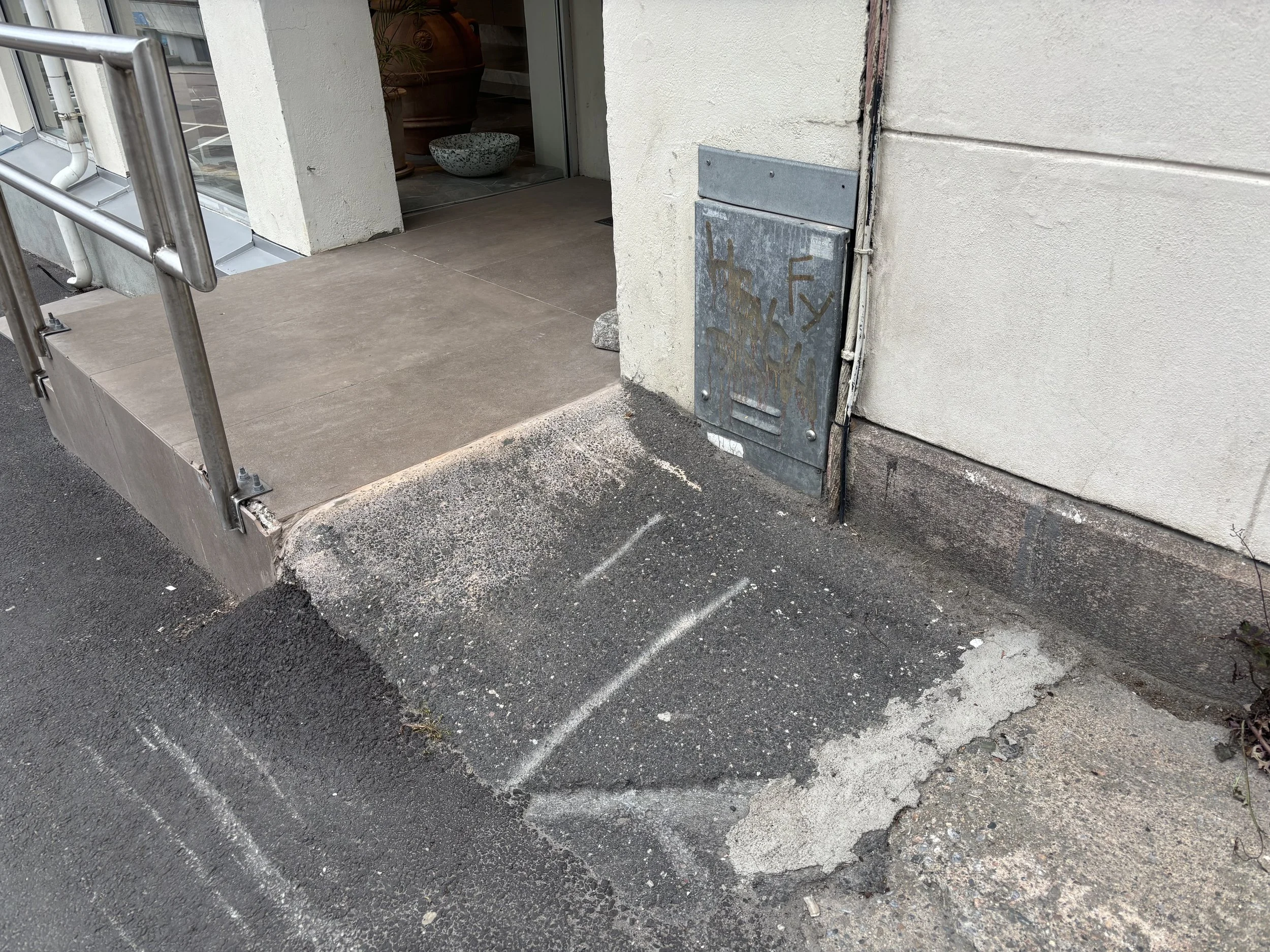 Nærbilde av asfaltklattens avslutning mot fortauet. Slitt, ujevn overflate og skrå avslutning på siden ned mot fortau, uten markering eller tydelig  overgang.