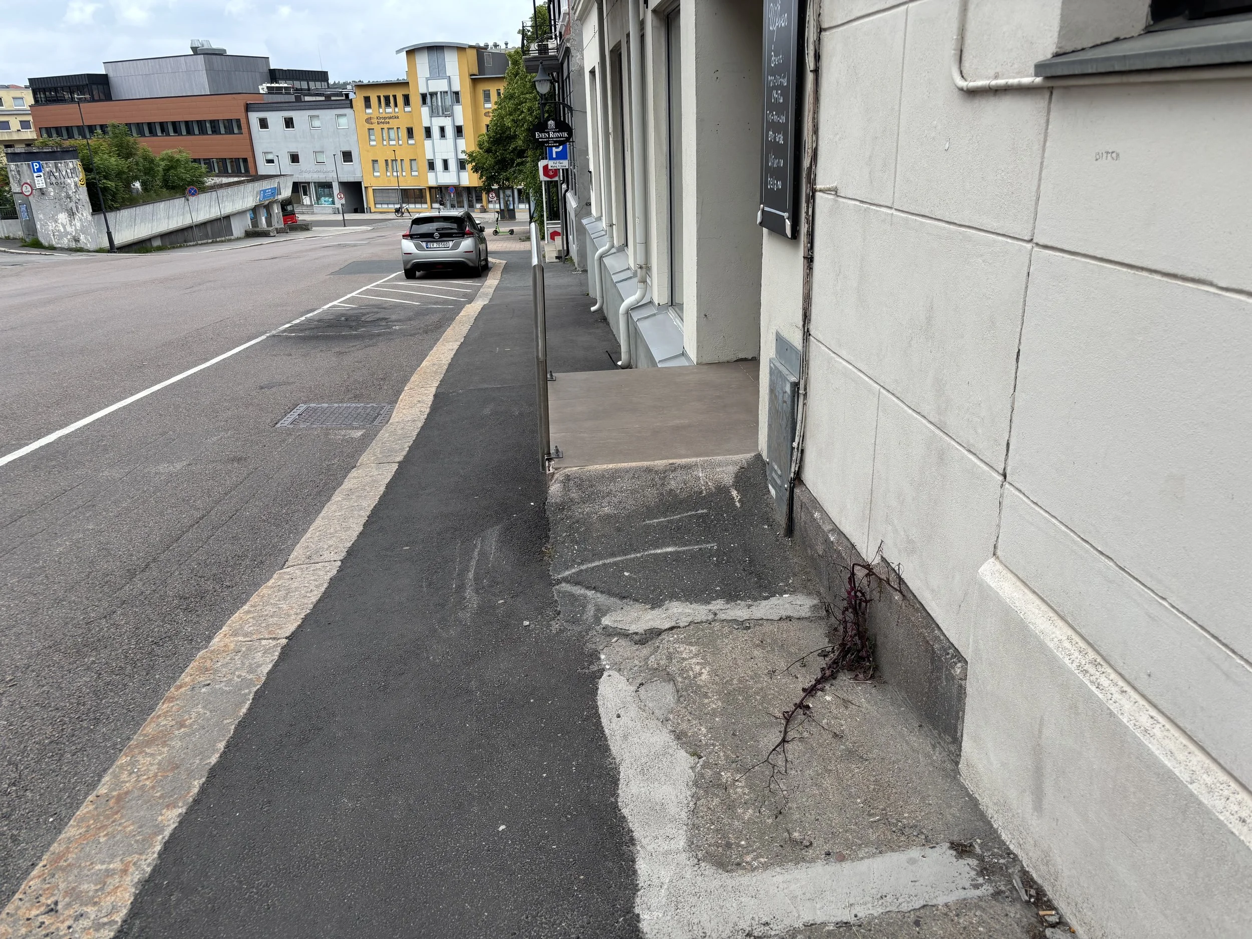 Oversiktsbilde som viser asfaltklattens tverrfall ned mot gaten, smal bredde , ujevn overflate og brå avslutning mot fortauet. Gelender synlig øverst på reposet.