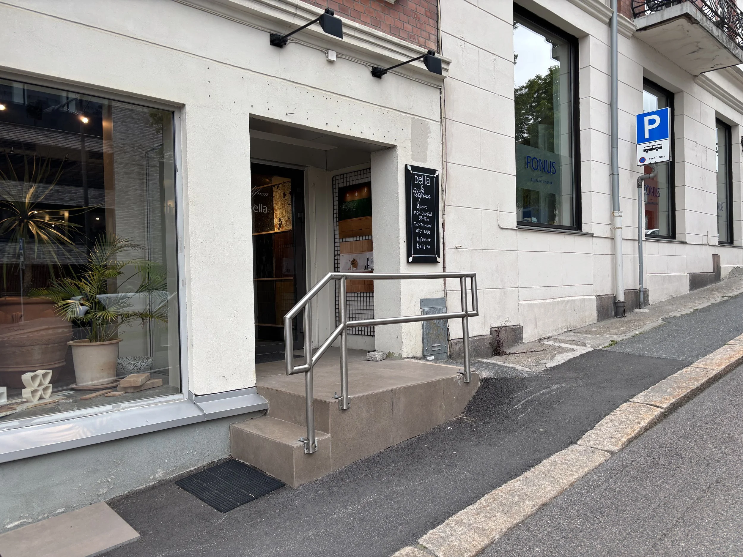 Fasade av eldre bygård med inngangsparti. En liten asfaltert rampe med gelender fører opp til et høyt trinn og inn i butikken.