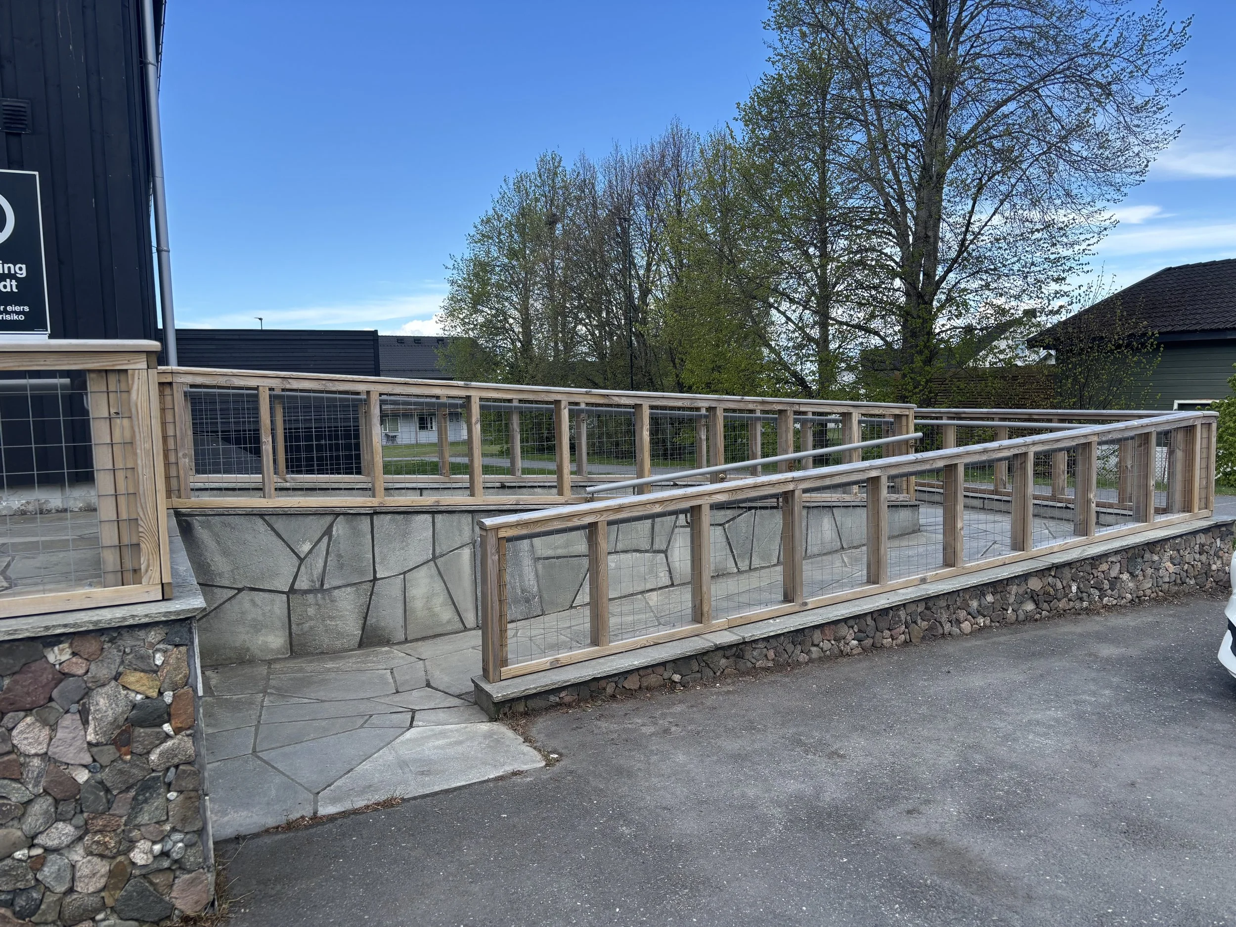 Rampe med trerekkverk, grovmasket netting og metallhåndløpere langs hele lengden, sett fra parkeringsplassen.