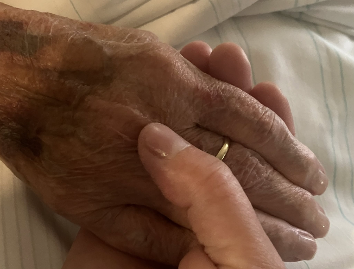Nærbilde av to hender som holder om hverandre. Den ene hånden er eldre med synlig aldret hud og gullring, den andre er yngre. Hendene hviler på et hvitt, stripete sengetøy.