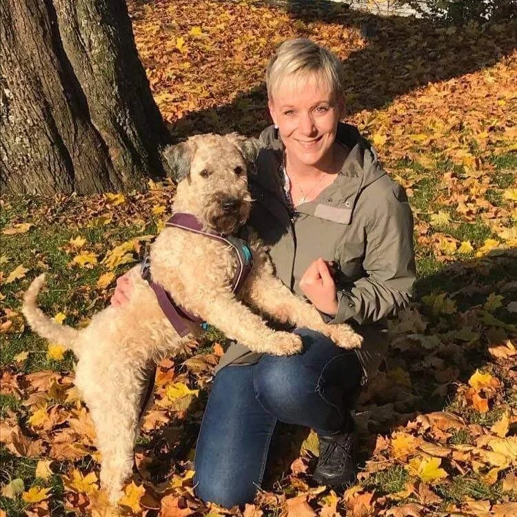 En kvinne med kort blondt hår smiler mens hun holder en liten hund med krøllete pels, omgitt av høstløv og trær.