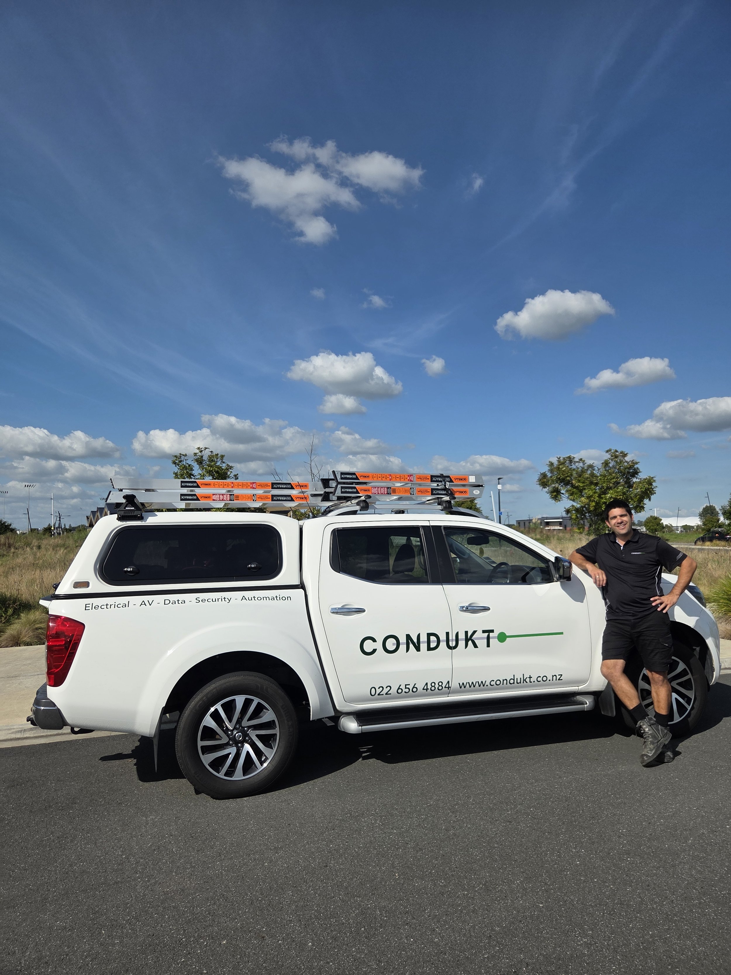 A man standing next to a white company vehicle branded with 'CONDUKT' in green letters, with a ladder on the roof and the company’s contact information. The man is smiling and has one hand on his hip, wearing a black shirt and shorts. The background features a clear blue sky with scattered clouds and some trees.
