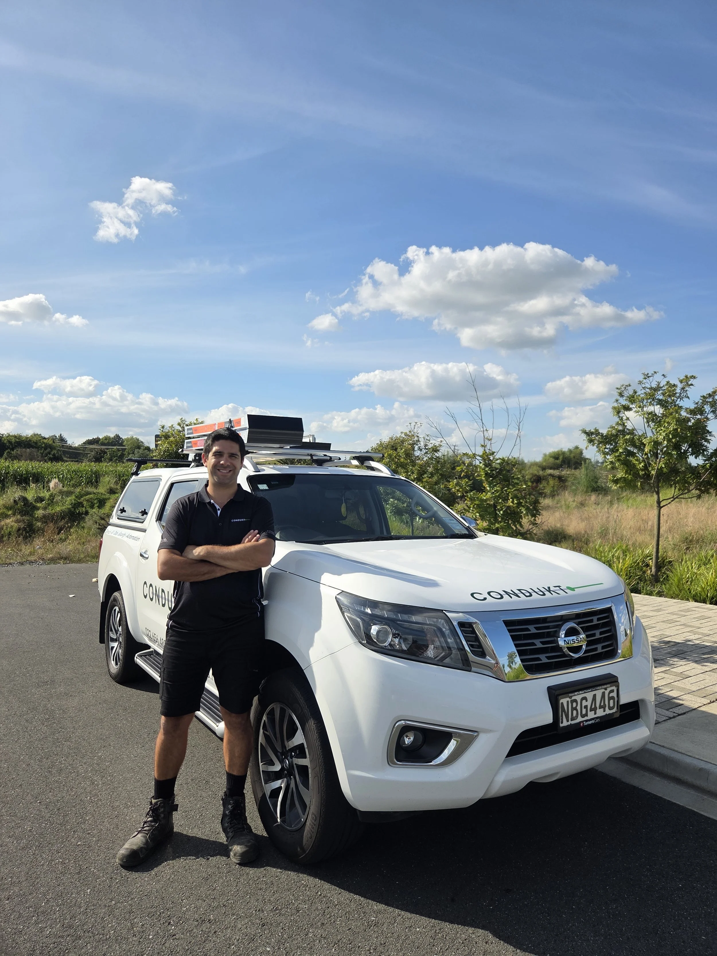 A man in black shorts and a black shirt stands with arms crossed next to a white Nissan vehicle with a logo that says 'CONDUKT' on the front.
