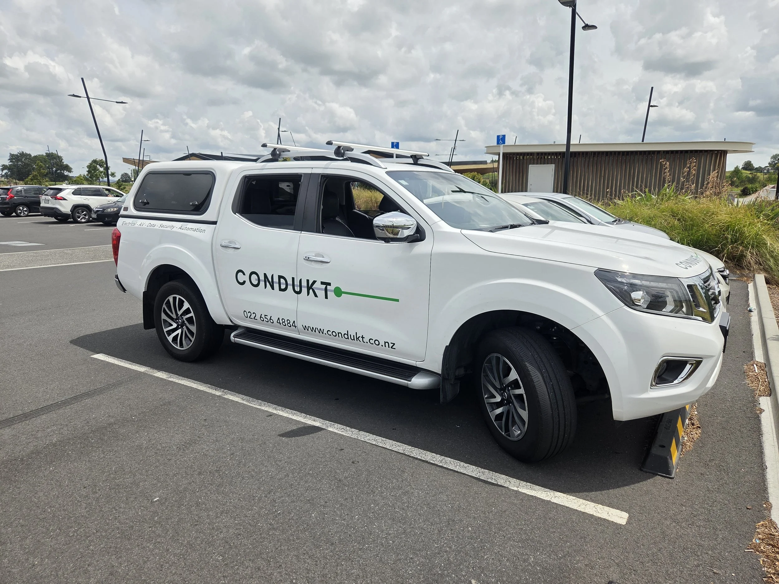 White vehicle with company branding parked in a lot, with other cars and a wooden building in the background.