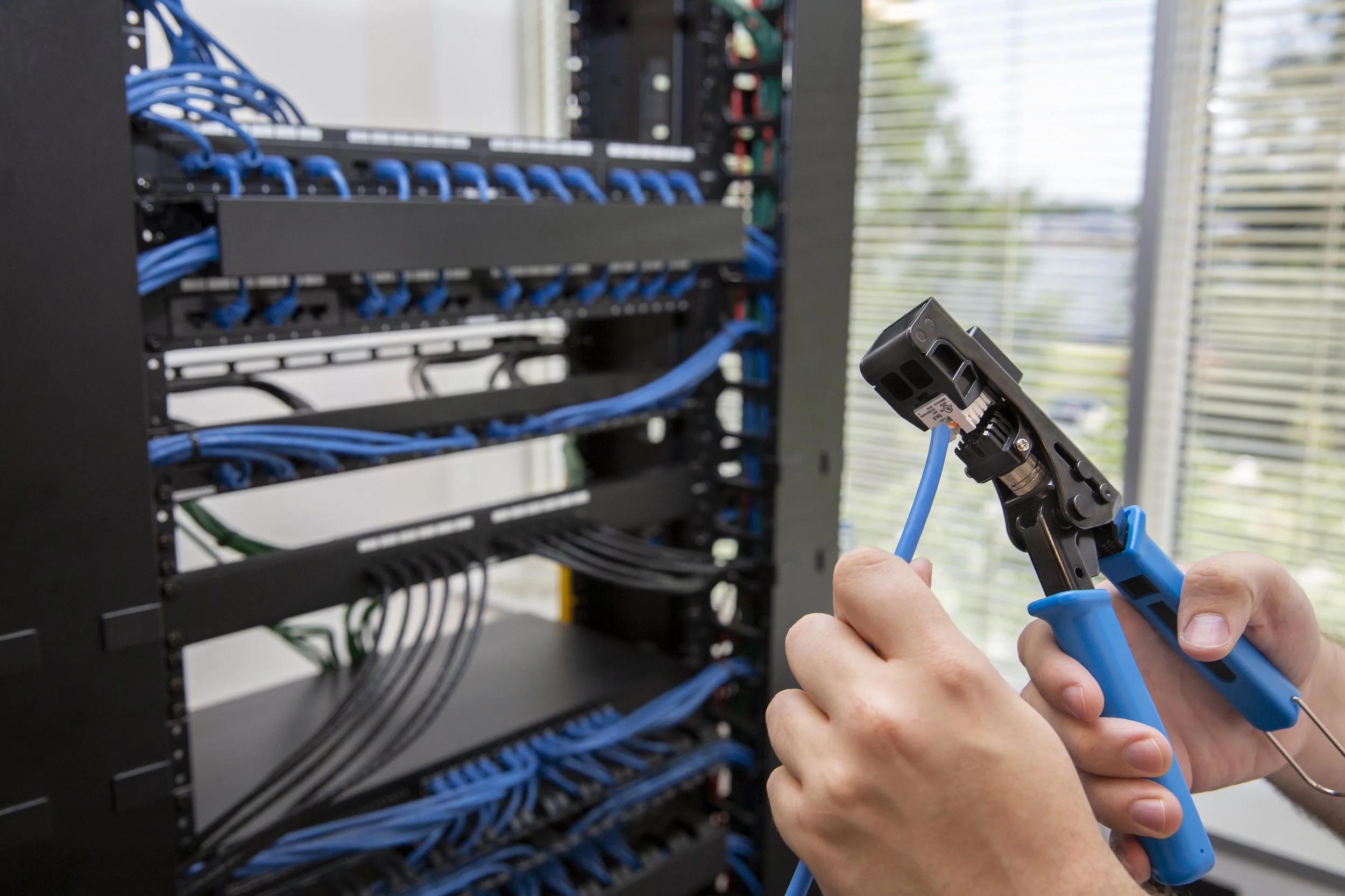 Person using crimping tool to connect blue Ethernet cable to a network patch panel with multiple blue cables in a server room.