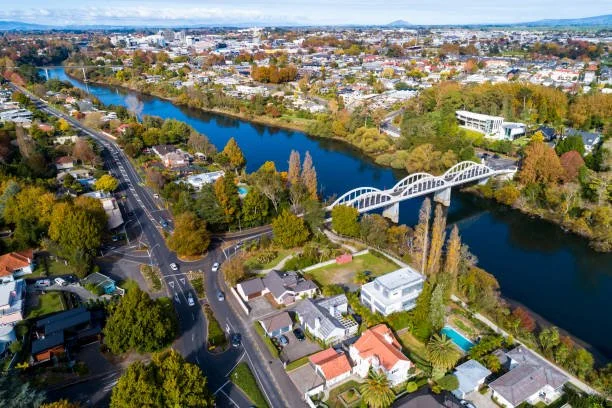 Aerial view of the Waikato river running through Hamilton City with a white bridge, surrounded by residential houses, trees, and roads in a suburban area.