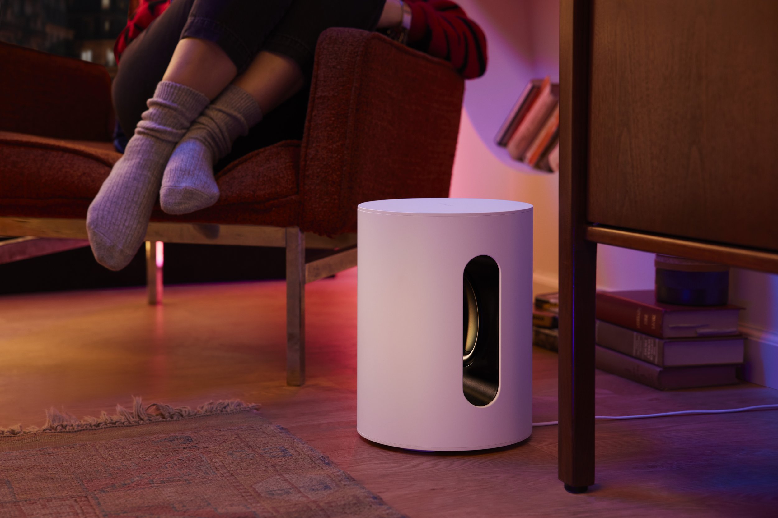 Modern white Sonos speaker with a circular black sound opening, placed on a wooden floor next to a bookshelf and a rug, in a cozy, dimly lit living room.
