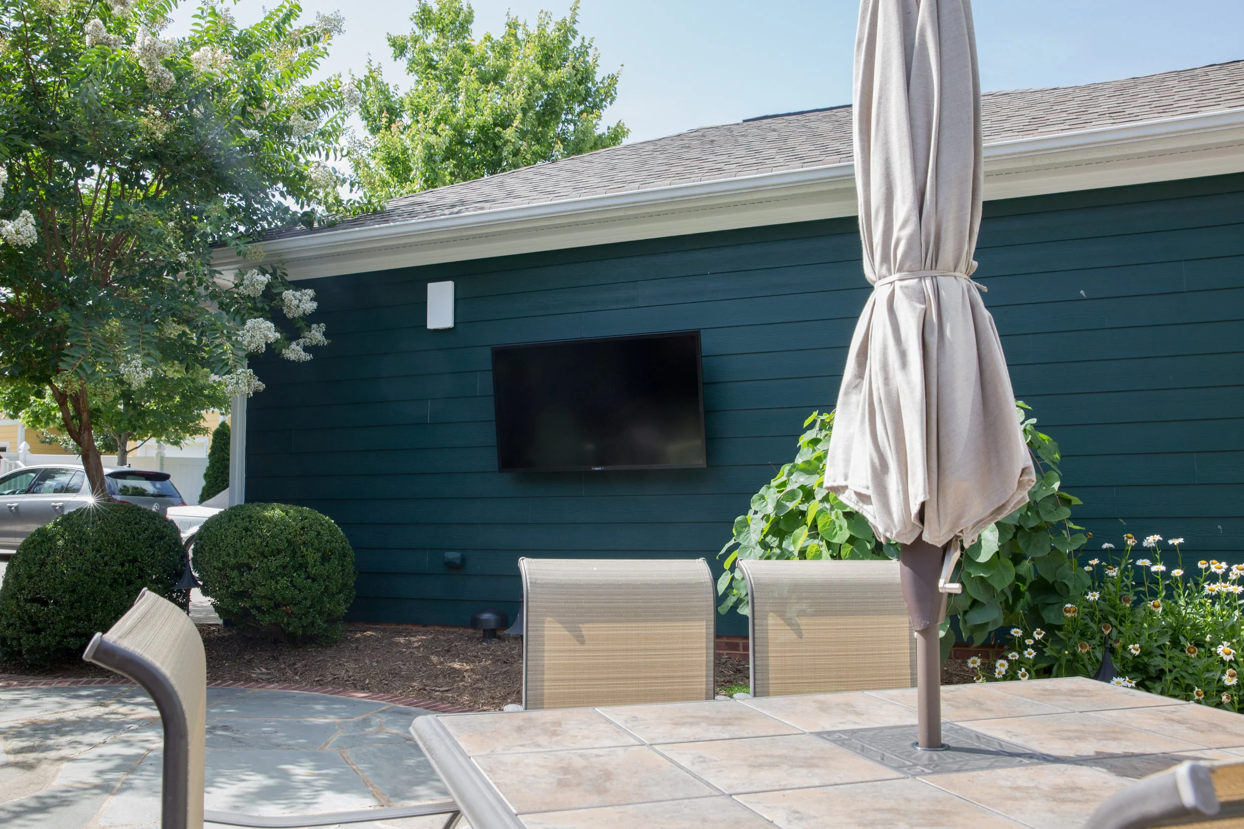 A backyard patio with a table, chairs, and a closed patio umbrella. A landscaped garden with bushes and flowers, a blue house wall with a mounted outdoor television, and a large tree with white blossoms are visible under a clear blue sky.