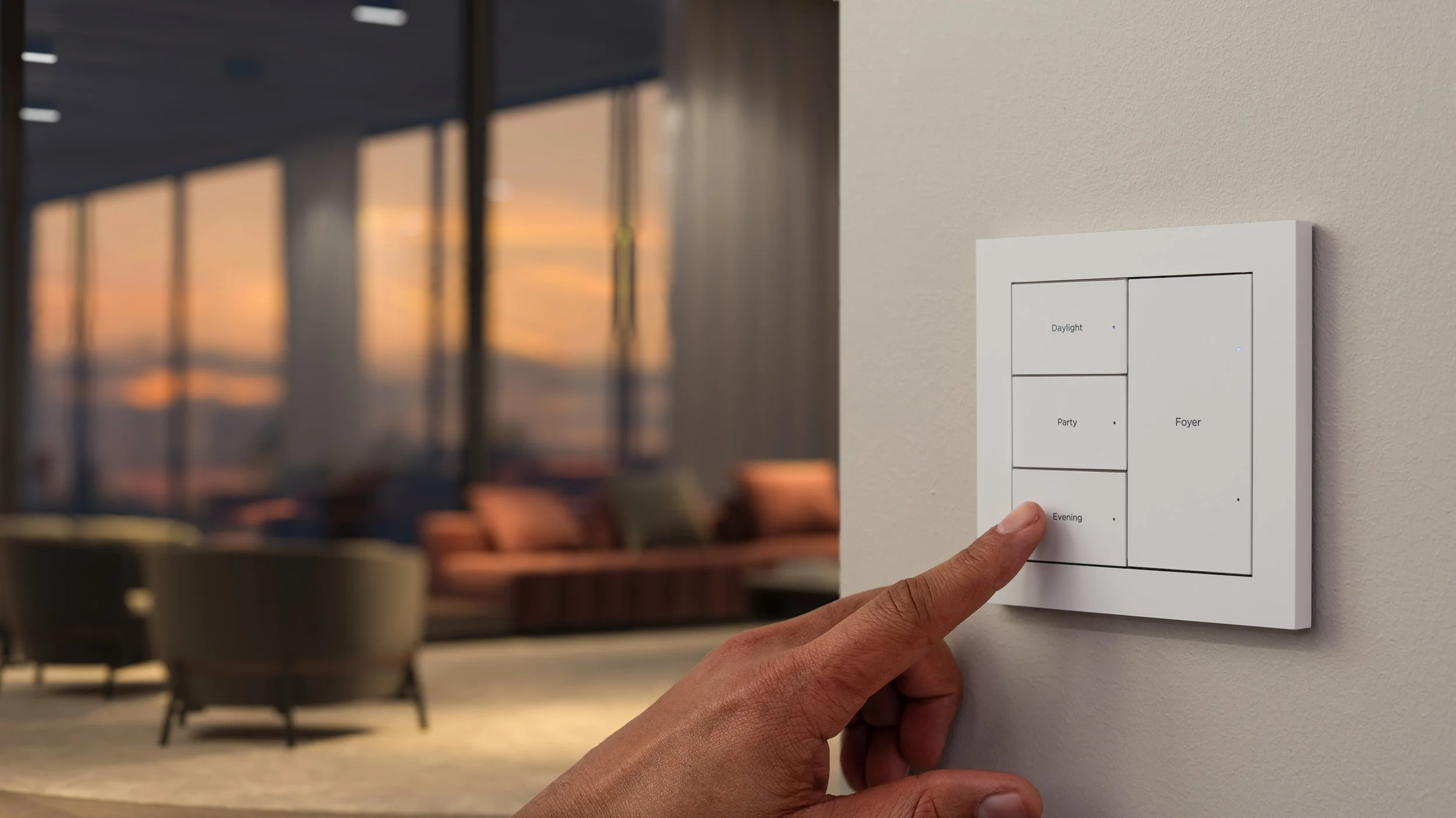 A person's hand pressing a button labeled 'Evening' on a white wall-mounted smart lighting control panel, with other buttons labeled 'Daylight,' 'Party,' and 'Foyer.' In the background, there is a blurred living room with large windows showing a sunset view, seating furniture, and cushions.