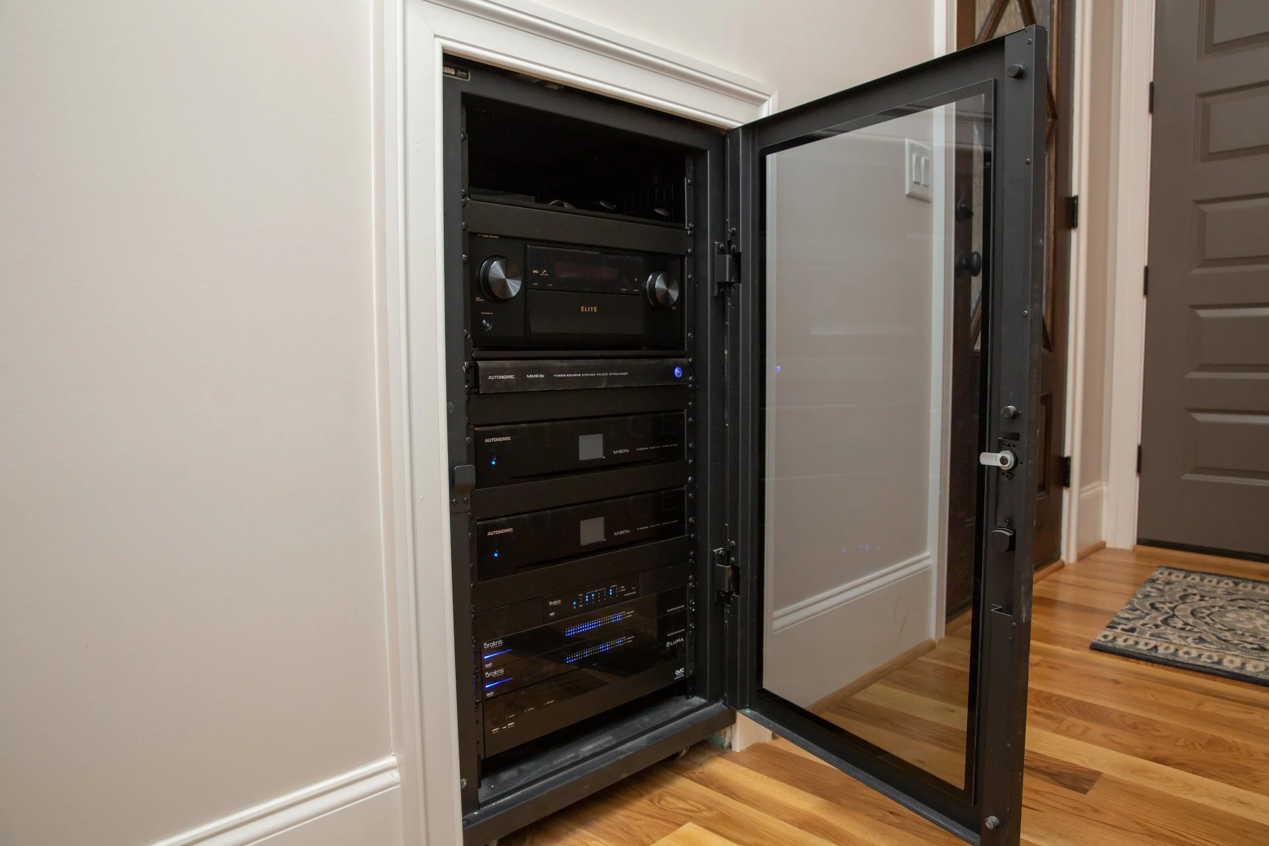 A black server rack with multiple electronic devices, including a receiver and other audio or network equipment, inside a room with wooden flooring and white walls. The rack door is open, revealing the equipment.