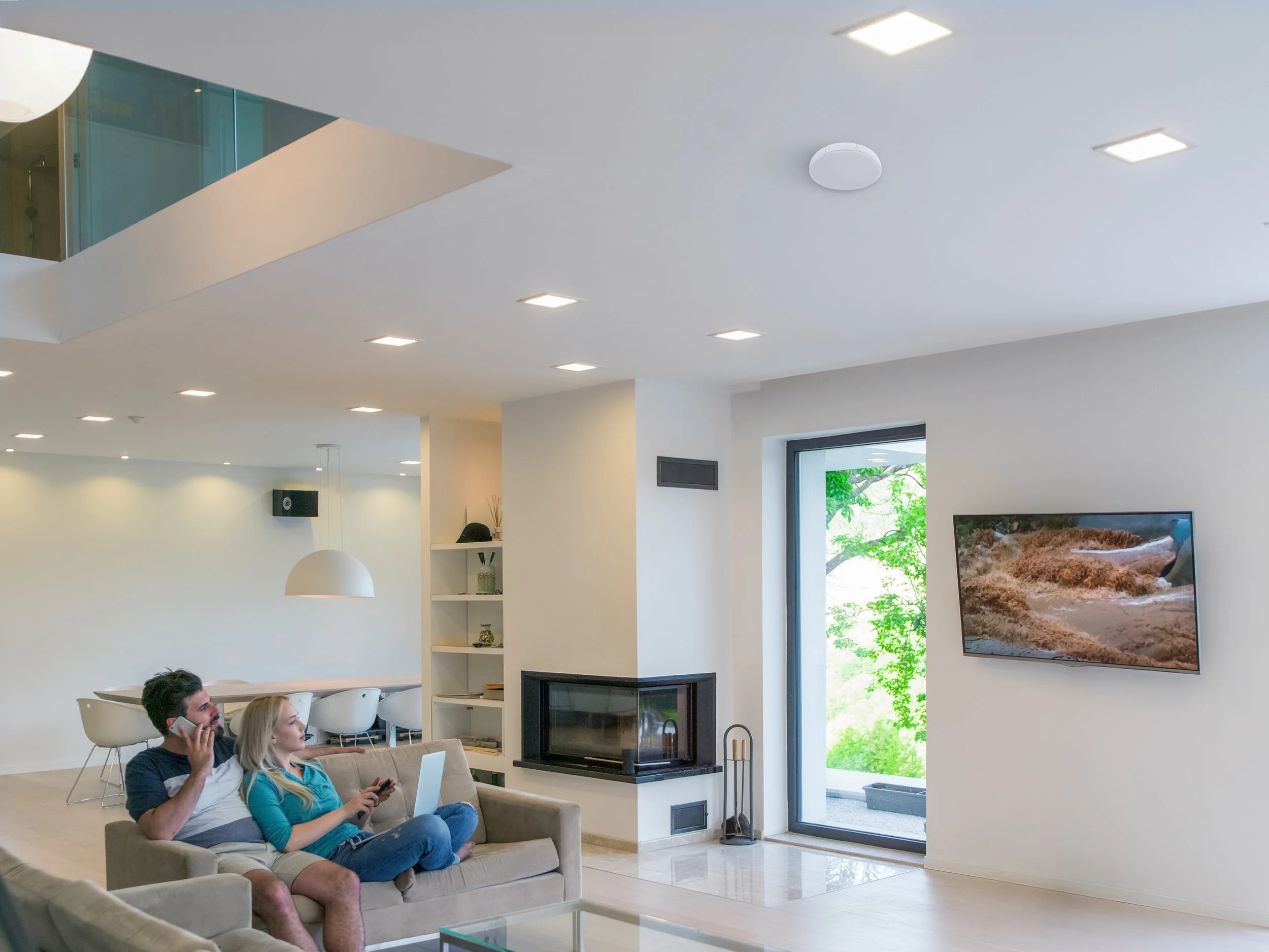 A modern living room with a beige sofa, two people sitting on it, a man talking on a phone, a woman using a tablet, a fireplace, a wall-mounted TV with nature scenery, a large window with greenery outside, and a staircase leading to an upper level with glass railing.
