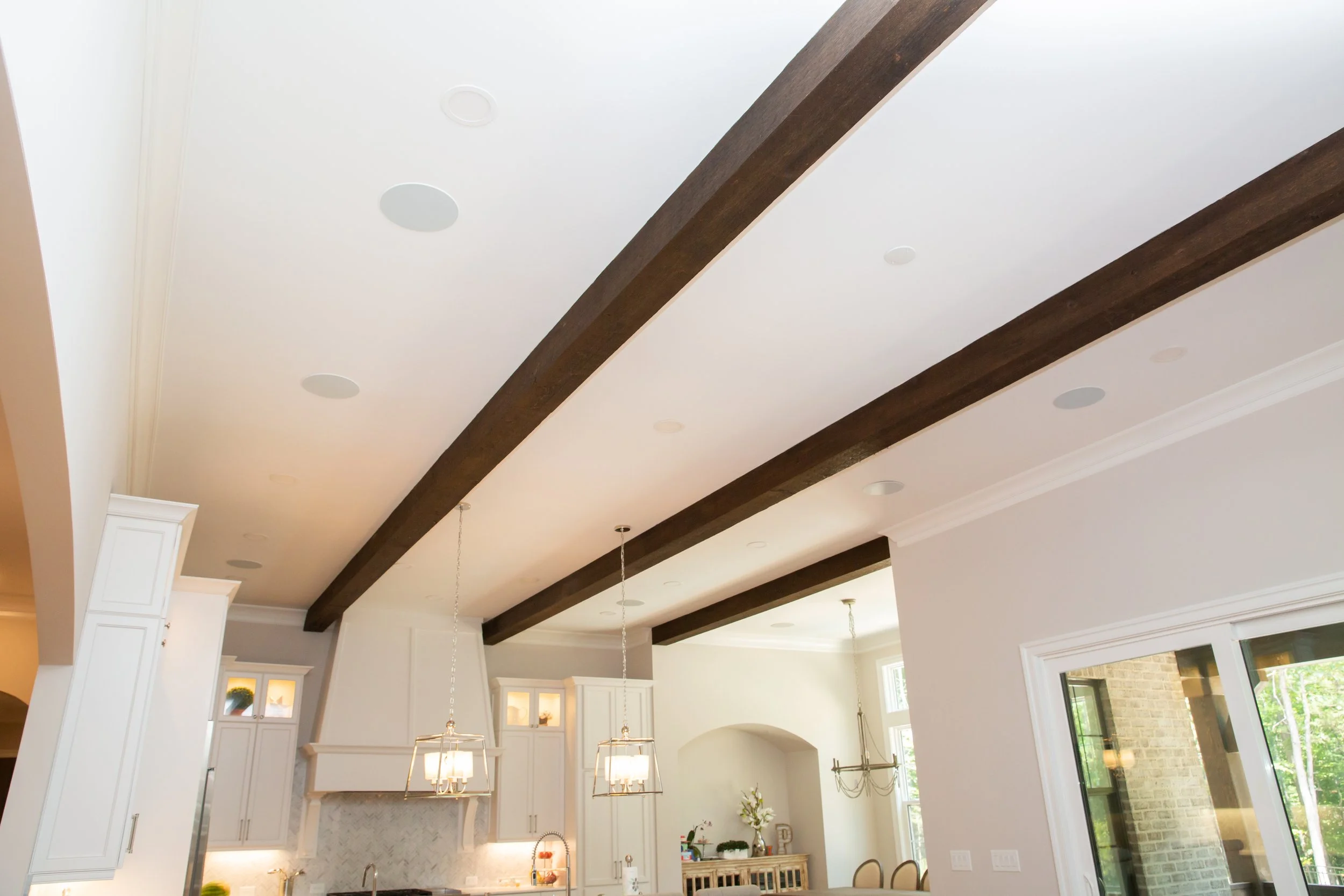 Open-concept kitchen and living area with exposed wooden beams and ceiling mount speakers, white cabinetry, and large windows letting in natural light.