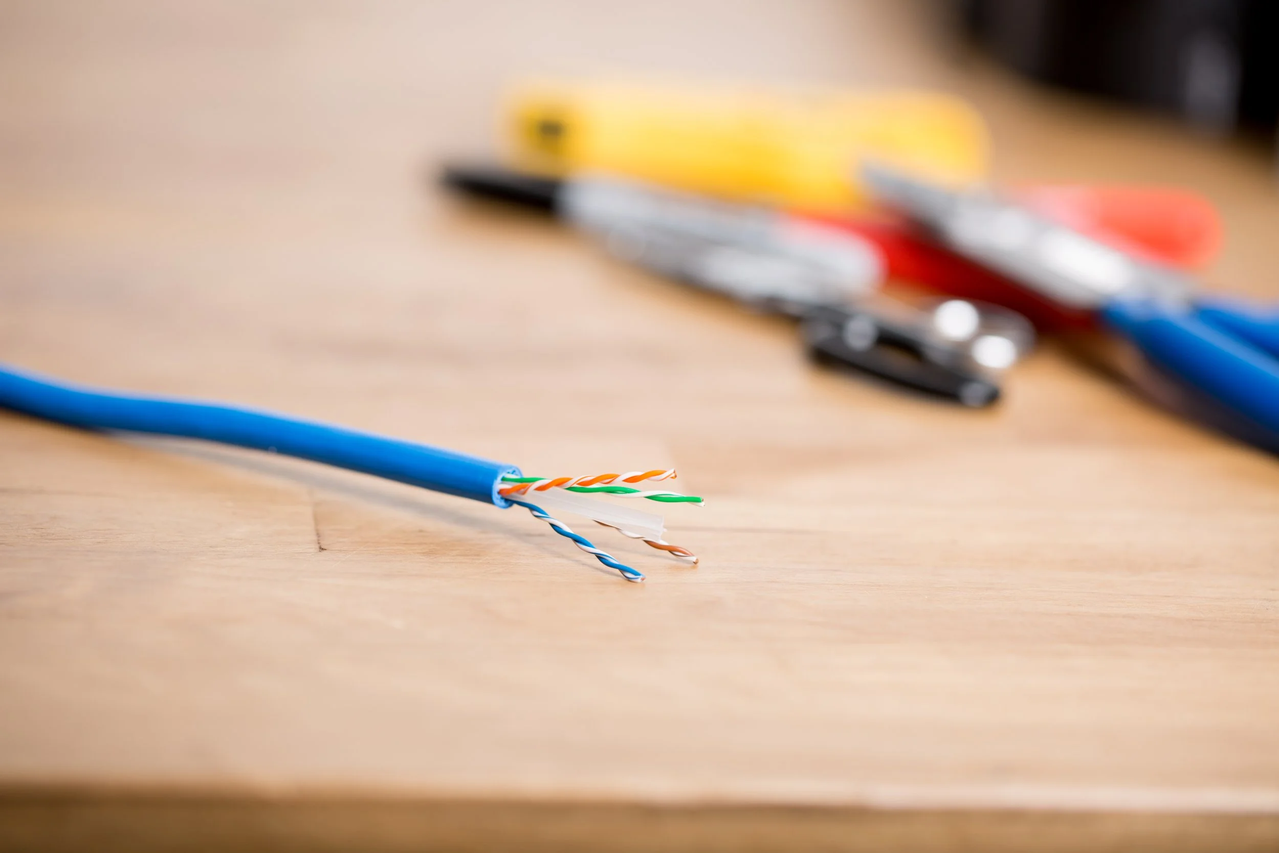 A close-up of a stripped blue Ethernet cable with exposed wires, lying on a wooden surface. In the background, out-of-focus tools such as scissors, electrical tape, and a utility knife are visible.
