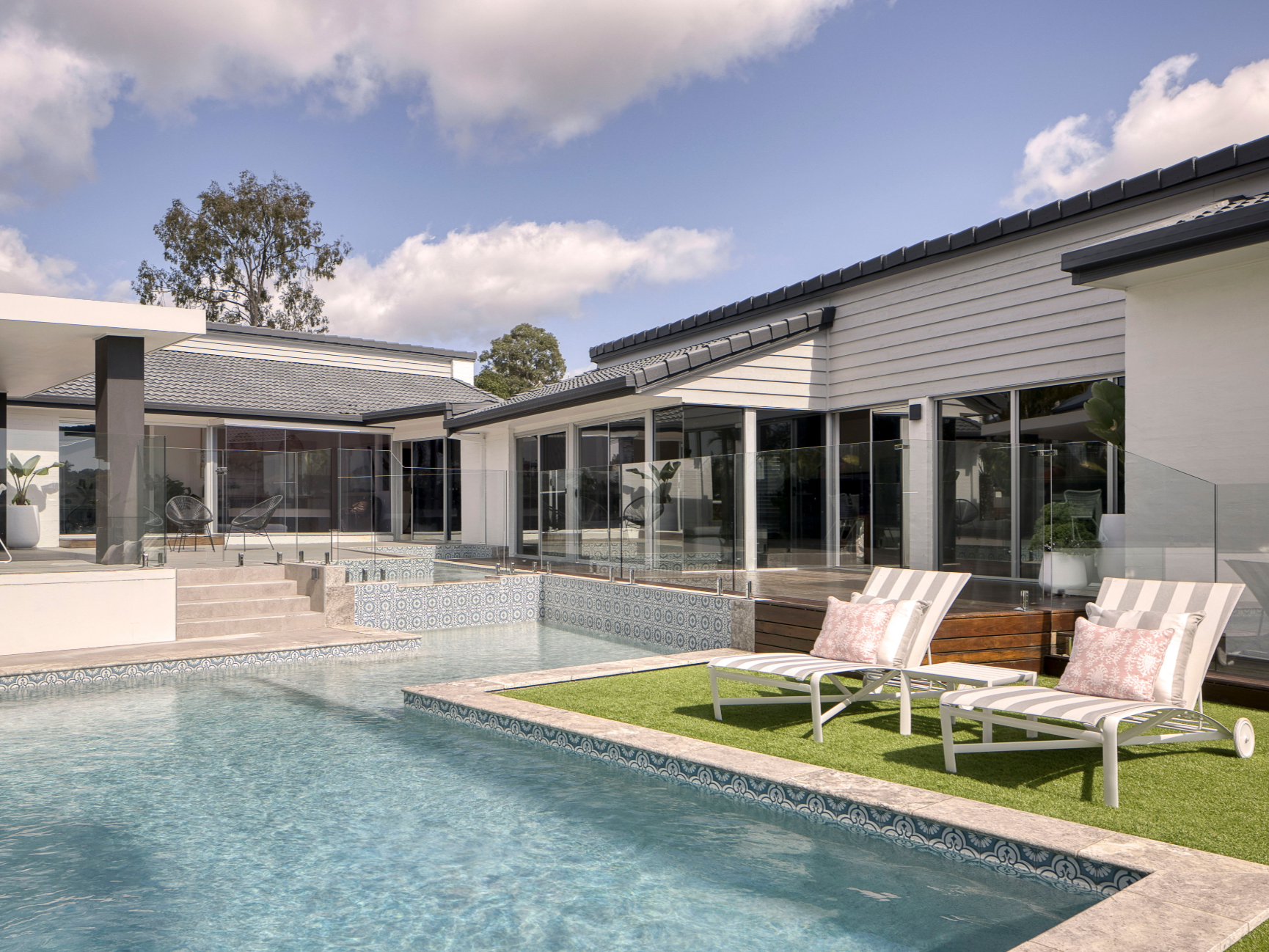 Luxury modern house with a swimming pool, lounge chairs with pink pillows, glass fencing, and patio area under a blue sky with clouds.