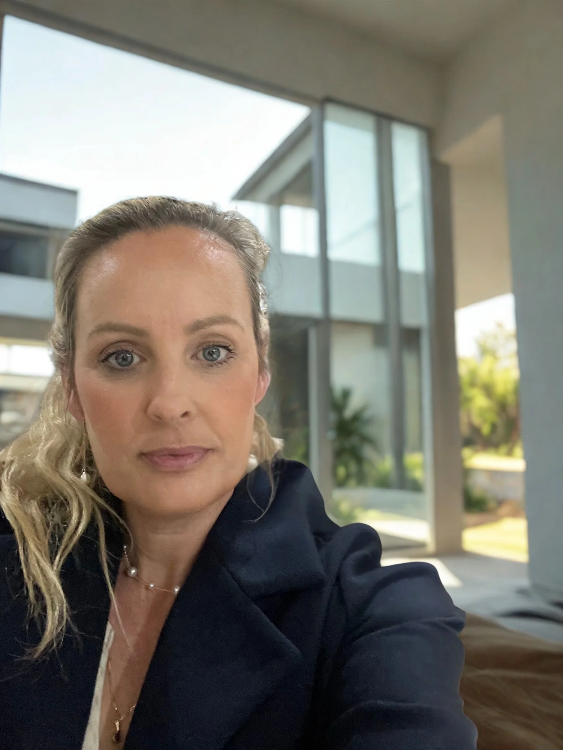 A woman with blonde hair and blue eyes taking a selfie in a modern house with large glass windows and a view of greenery outside.