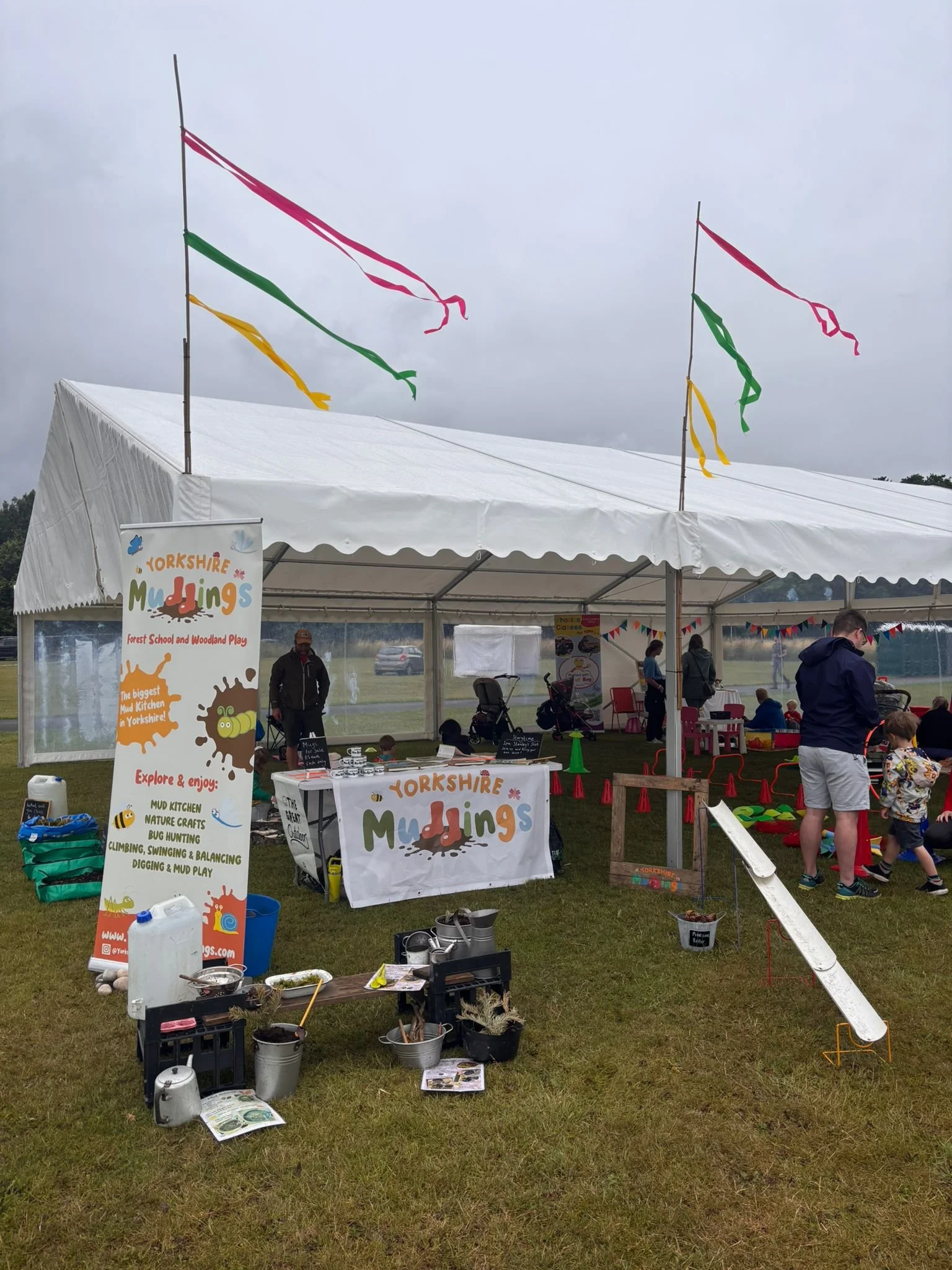 A white event tent with colorful flags on top, set up outdoors on a cloudy day. Inside, there are people, children, tables, and activities related to Yorkshire mudding, a forest school and woodland play event. There are signs advertising mud kitchens, nature crafts, bug hunting, climbing, swinging, balancing, digging, and mud play.