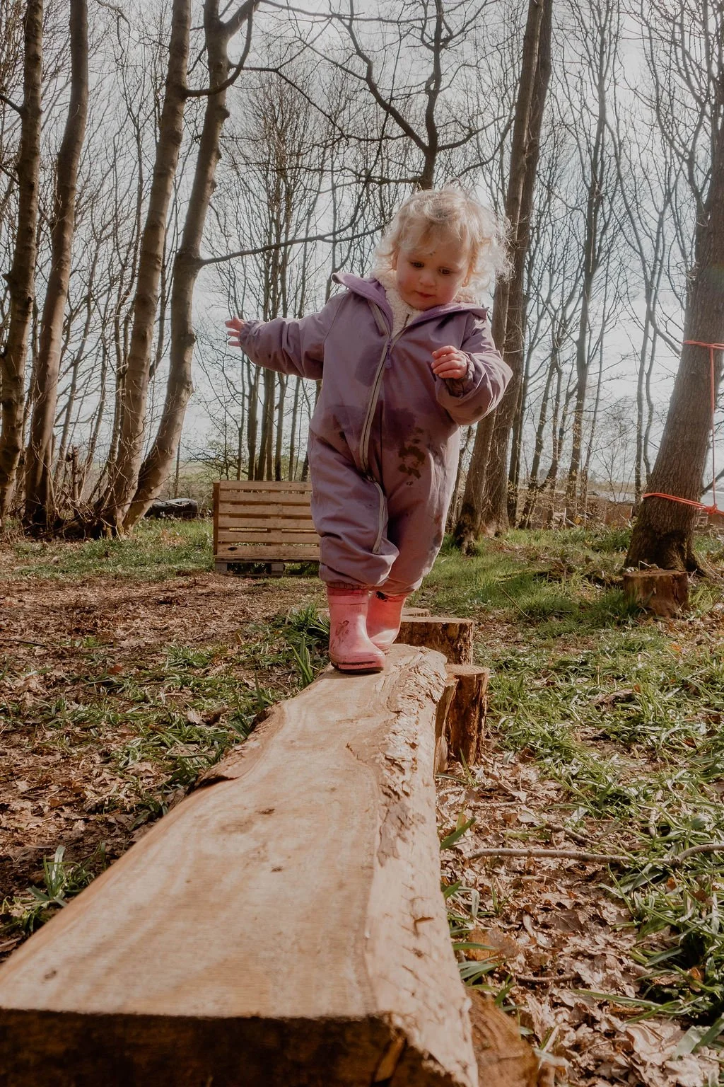 A young girl with curly blonde hair wearing a purple jumpsuit and pink boots balancing on a wooden log in a forested area.