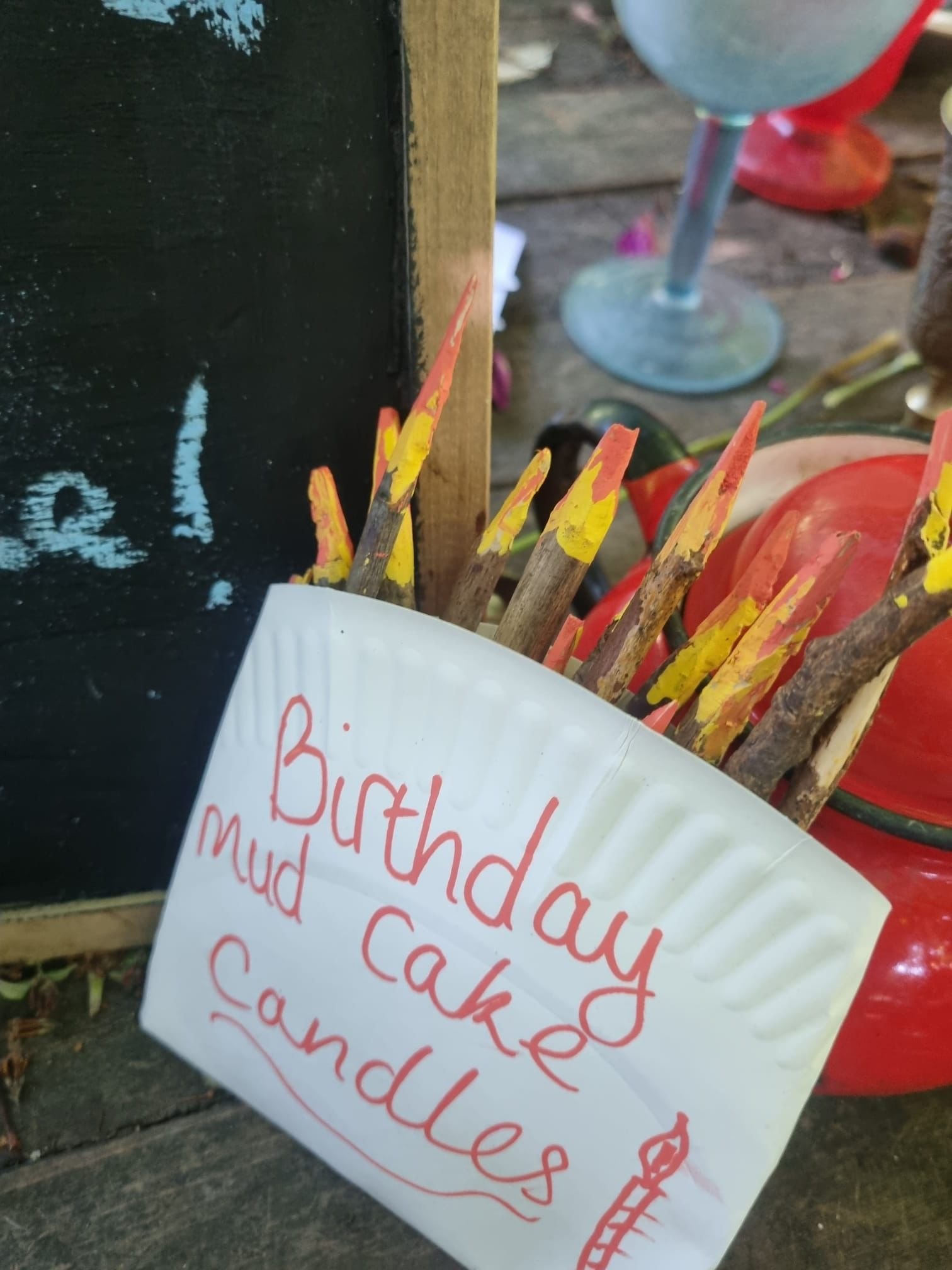 A sign that reads 'Birthday mud cake candles' and a collection of colorful paintbrushes with yellow and orange paint on the bristles, placed on a wooden surface.