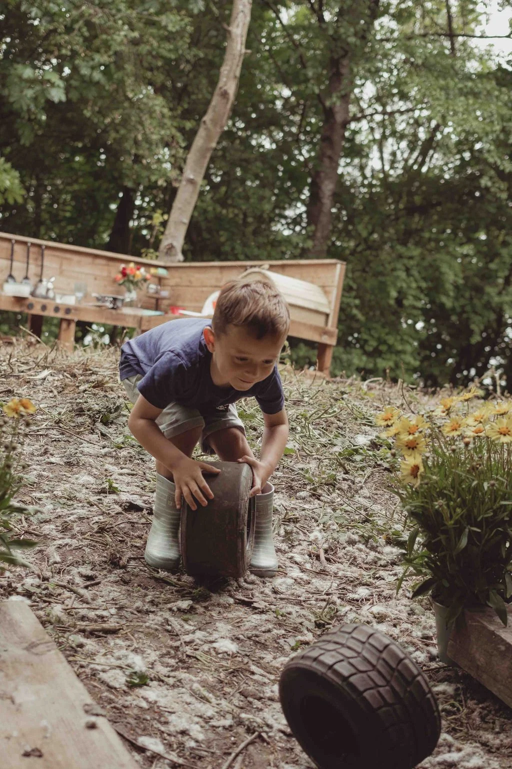 A young boy in a blue shirt and shorts is crouched on the ground playing with a tire, using it as a pretend object, surrounded by garden and outdoor elements like flowers, trees, and a wooden deck with pots and gardening tools.