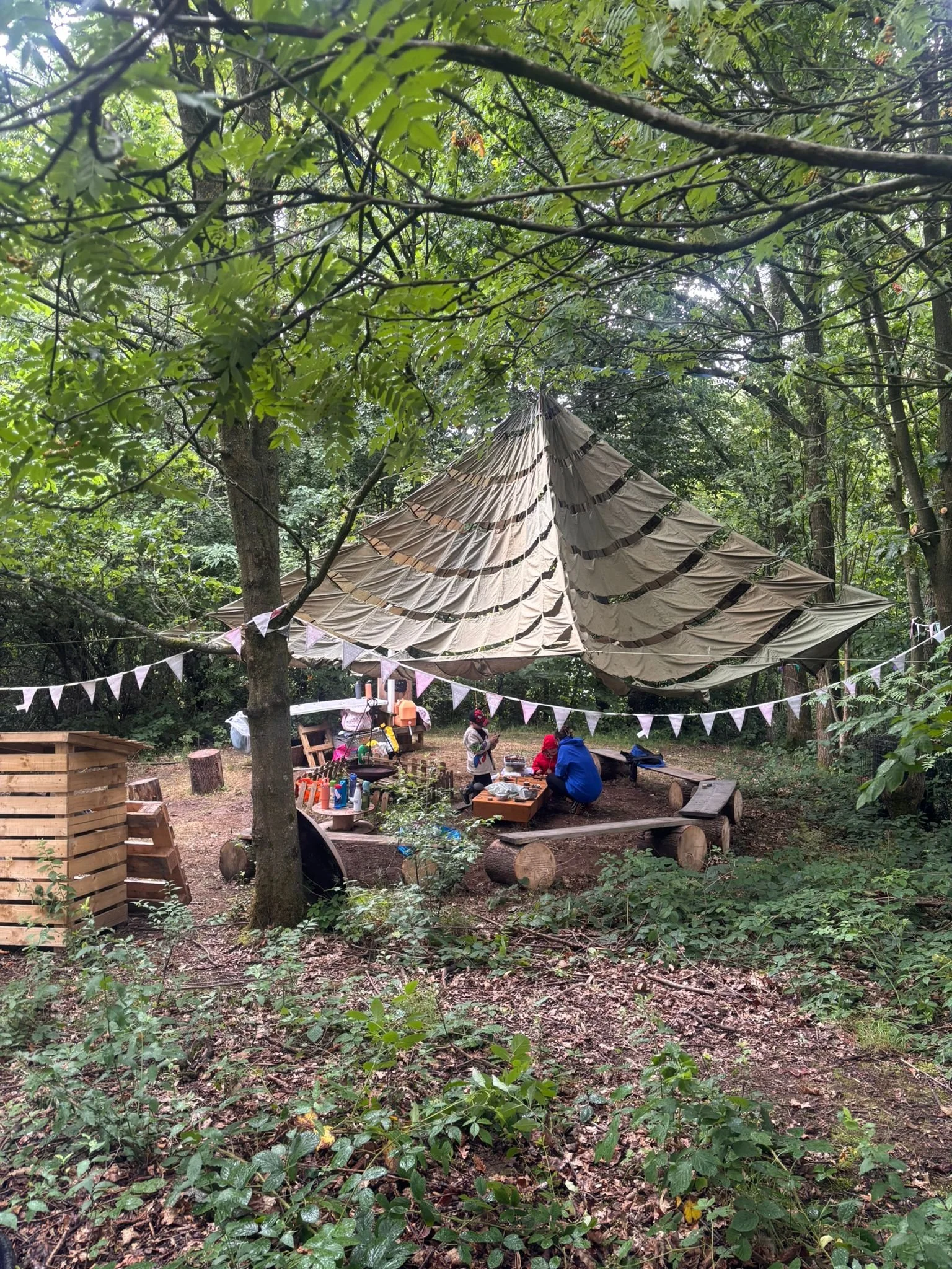 People sitting around a table under a large tarp canopy in a forest clearing, with bunting flags hanging overhead and wooden benches and stools around.
