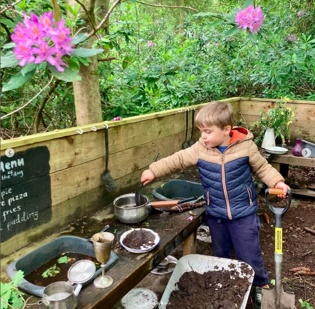 A young boy with short blonde hair wearing a tan and blue jacket and dark pants, gardening in an outdoor space. He is holding a small shovel, digging into dark soil, with a wooden fence and lush greenery with purple flowers in the background. The yard has various gardening tools and containers, including a watering can and a black tray with soil, on a wooden work surface.