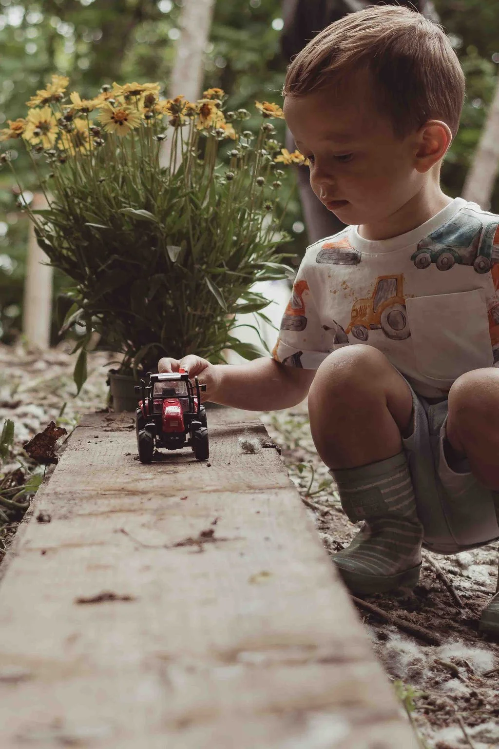 Young boy playing with a toy tractor outdoors, kneeling on the ground next to a wooden plank, with a potted plant and trees in the background.