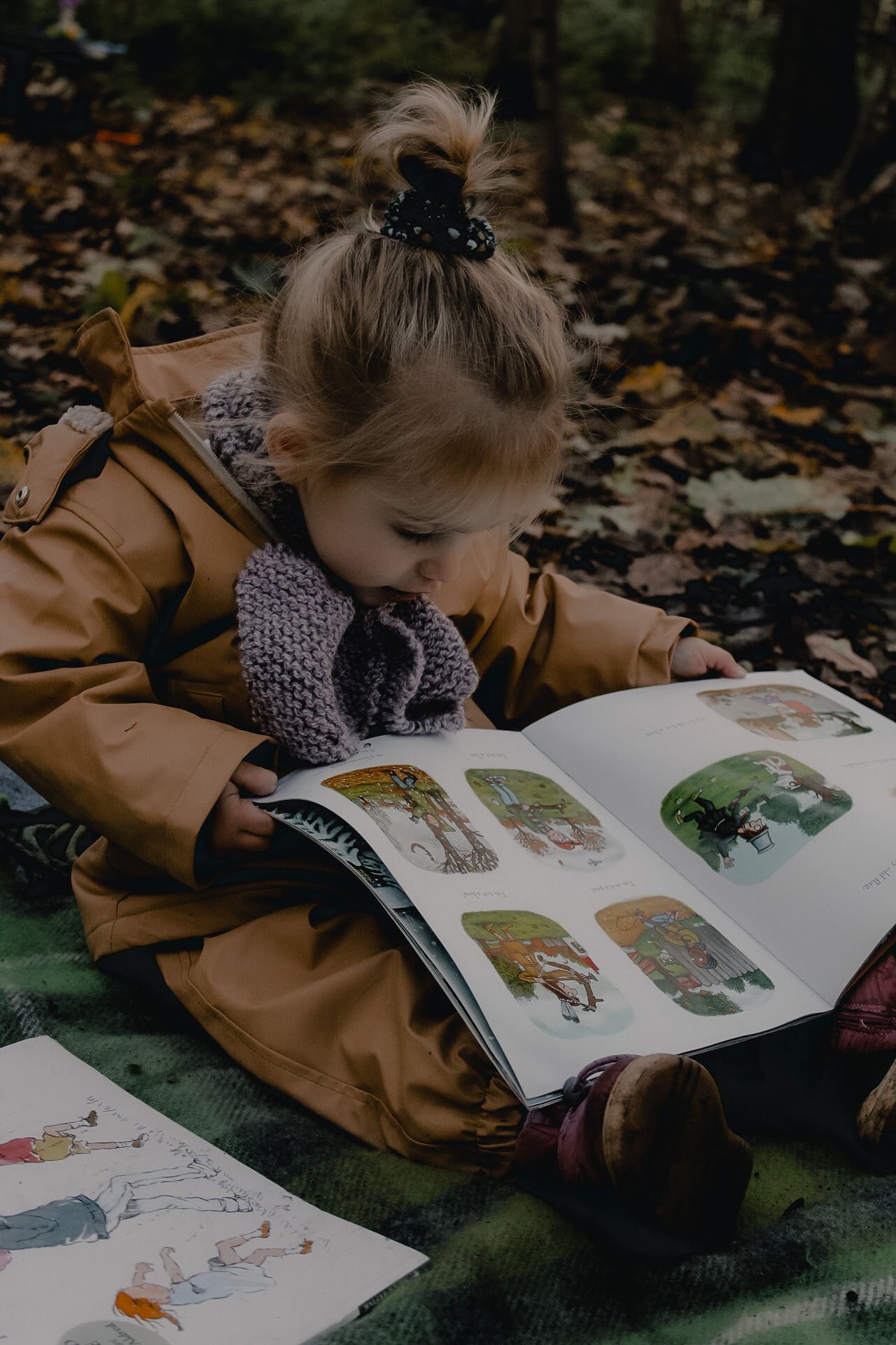 A young girl with a high ponytail tied with a black scrunchie, wearing a tan jacket and cozy scarf, sitting outdoors on a green blanket, reading a colorful children's storybook among fallen autumn leaves.