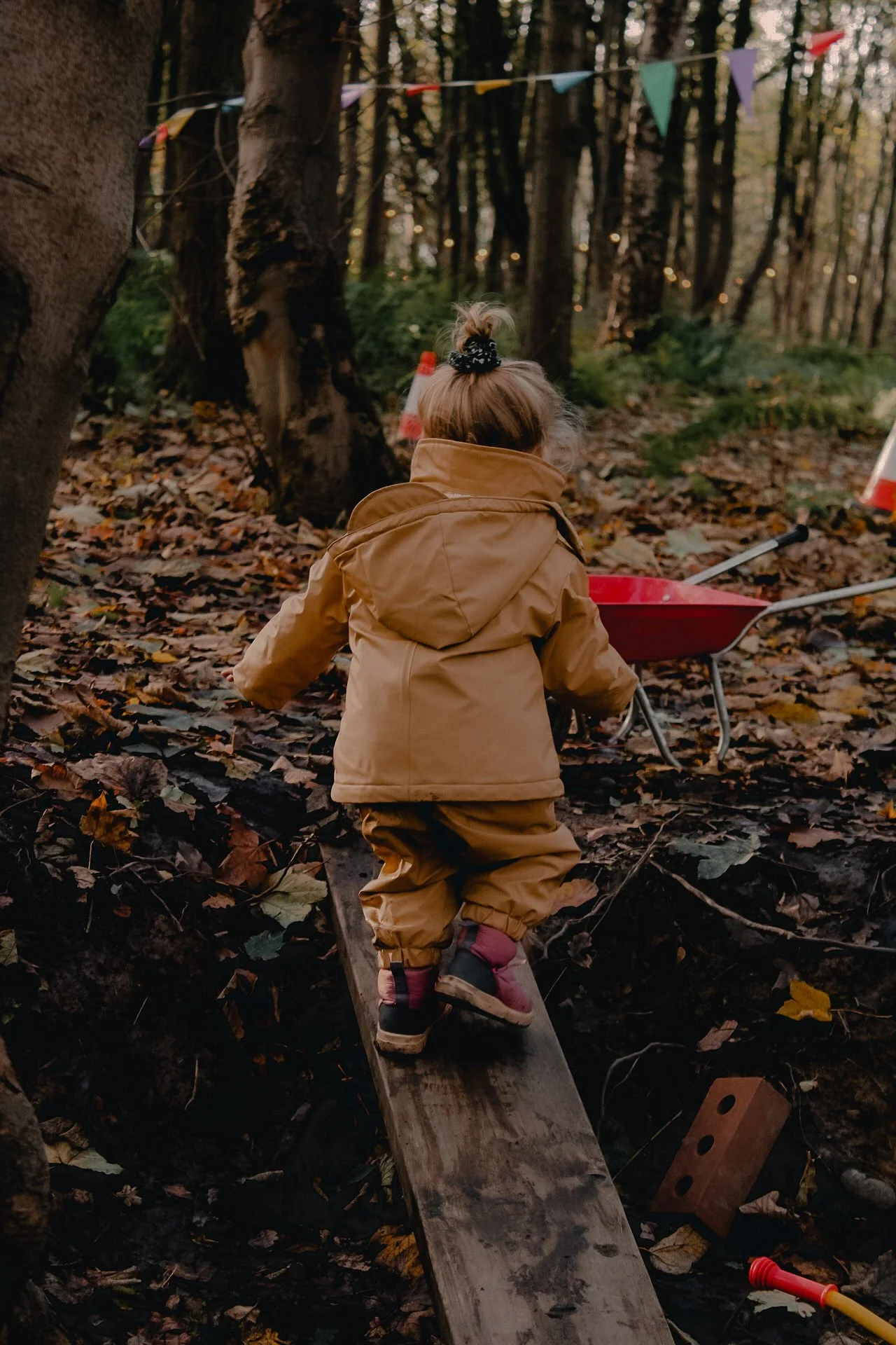 A young child in tan outdoor clothing and pink boots walking on a wooden plank over a forest floor covered in leaves, with a red wheelbarrow and colorful triangular flags in the background.