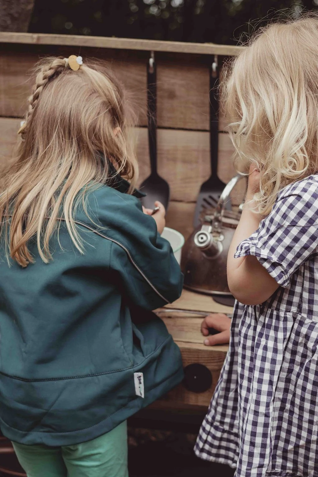 Two young girls with light blond hair looking at cooking utensils hanging on a wooden wall.