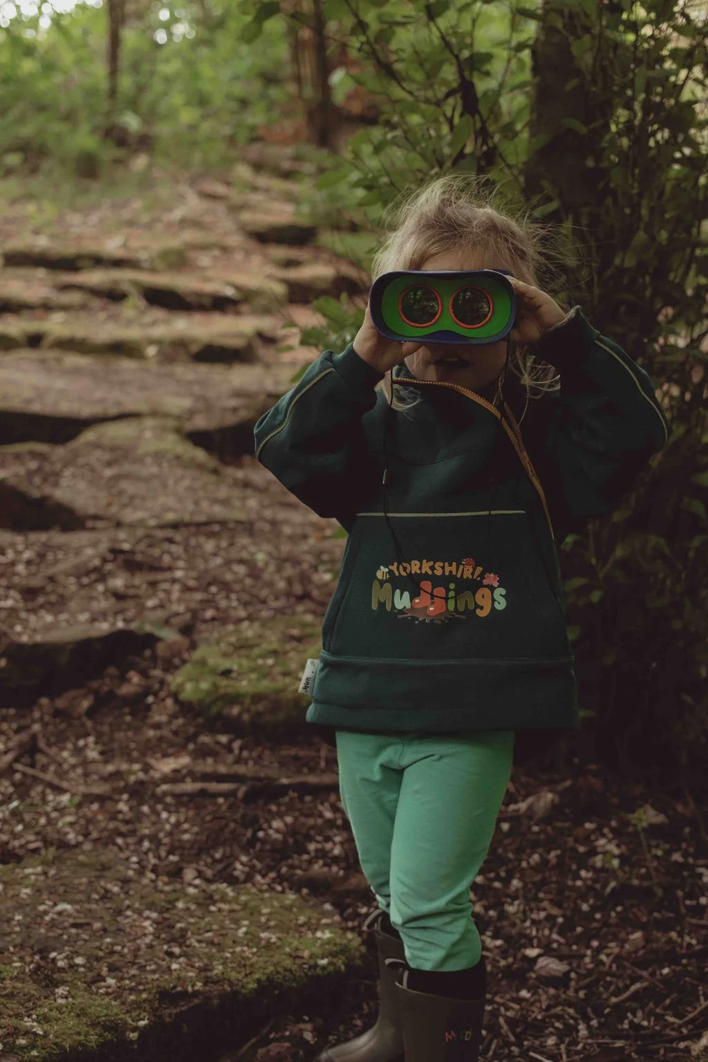 A young girl standing on a forest trail, holding a toy or device with two circular lenses up to her eyes, wearing a dark jacket with colorful text and light green pants.
