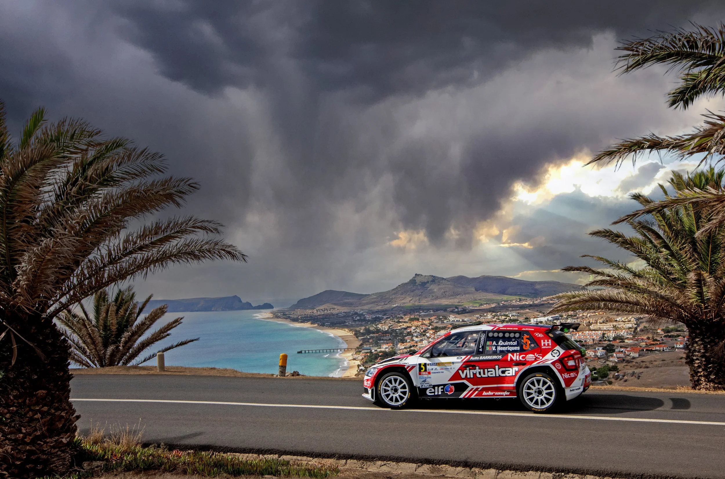 A rally car driven by A. Quintal and V. Henriques on a coastal road with palm trees, overlooking a beach and mountain landscape under dark, stormy clouds.
