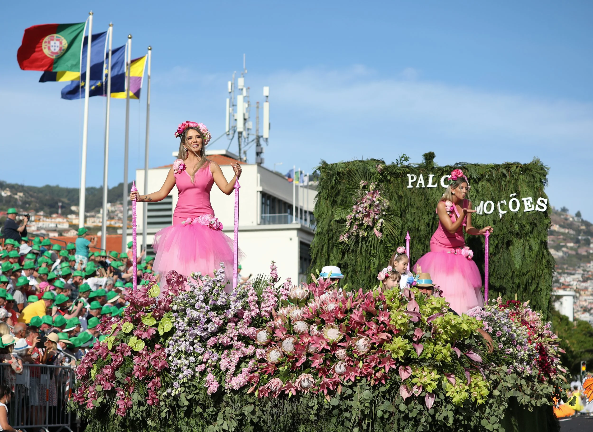 A parade float decorated with pink flowers and greenery, with women in pink dresses on top, one smiling and holding a pink scepter, and another reading a card. There is a crowd of spectators with green hats and flags in the background.